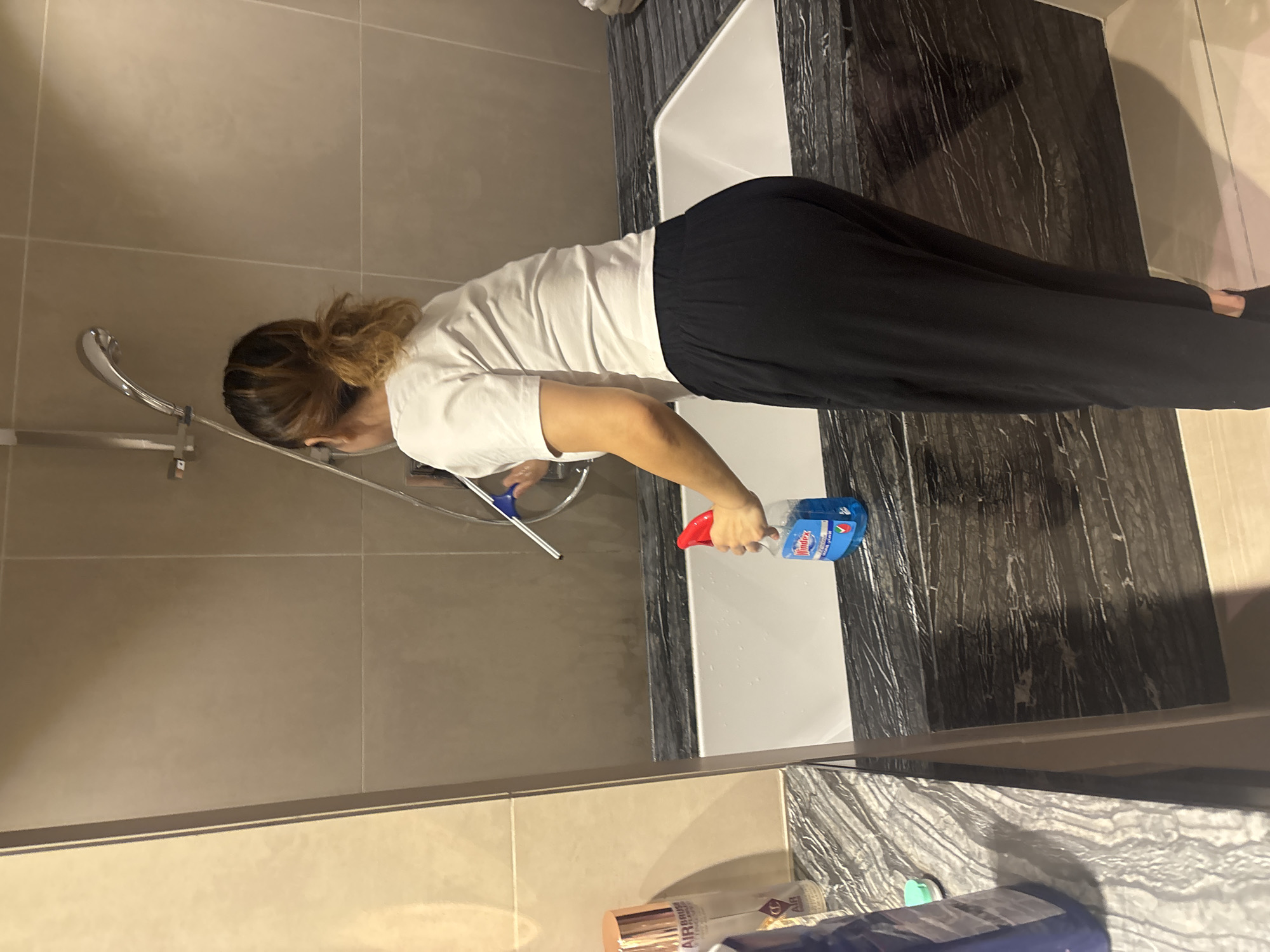 A woman with dark hair tied in a bun is cleaning a bathtub with a spray bottle and a squeegee. She is wearing a white t-shirt and black pants. The bathtub is white and surrounded by a dark marble countertop. In the background, there are beige tiled walls and a showerhead. On the countertop, there are several cleaning products and bottles. The scene appears to be in a modern bathroom.