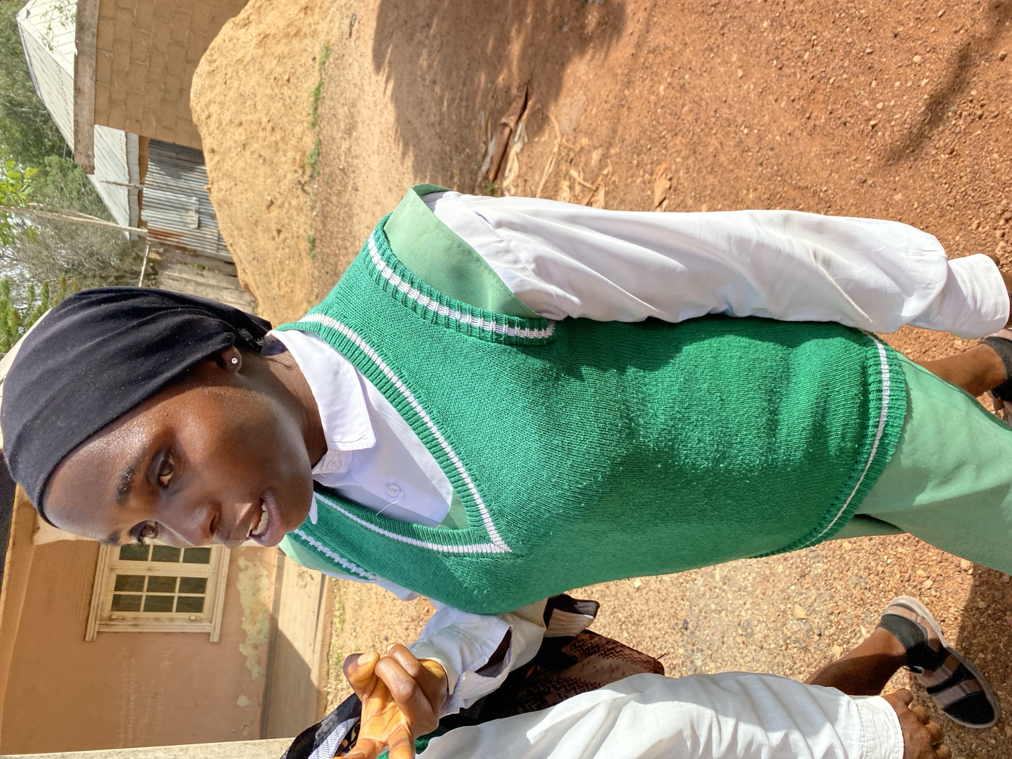 In Jos, Nigeria, a young person wearing a black hijab, white shirt, and a green knitted vest with white stripes is the main subject. They are looking towards the left, with an engaged expression and their mouth slightly open. Behind them, a simple building with a small, multi-paned window is visible. The building's walls appear to be a muted brown or pinkish color with some peeling plaster.
The surrounding environment is earthy and dry, with reddish-brown soil and small stones. In the background, there's a glimpse of a corrugated metal roof on another structure and a large, sandy mound. The overall scene suggests an outdoor setting, possibly a schoolyard or a rural area, under bright daylight. The lighting indicates it is daytime, with shadows cast on the ground, suggesting a sunny environment. The person's attire, particularly the vest, hints at a school uniform.