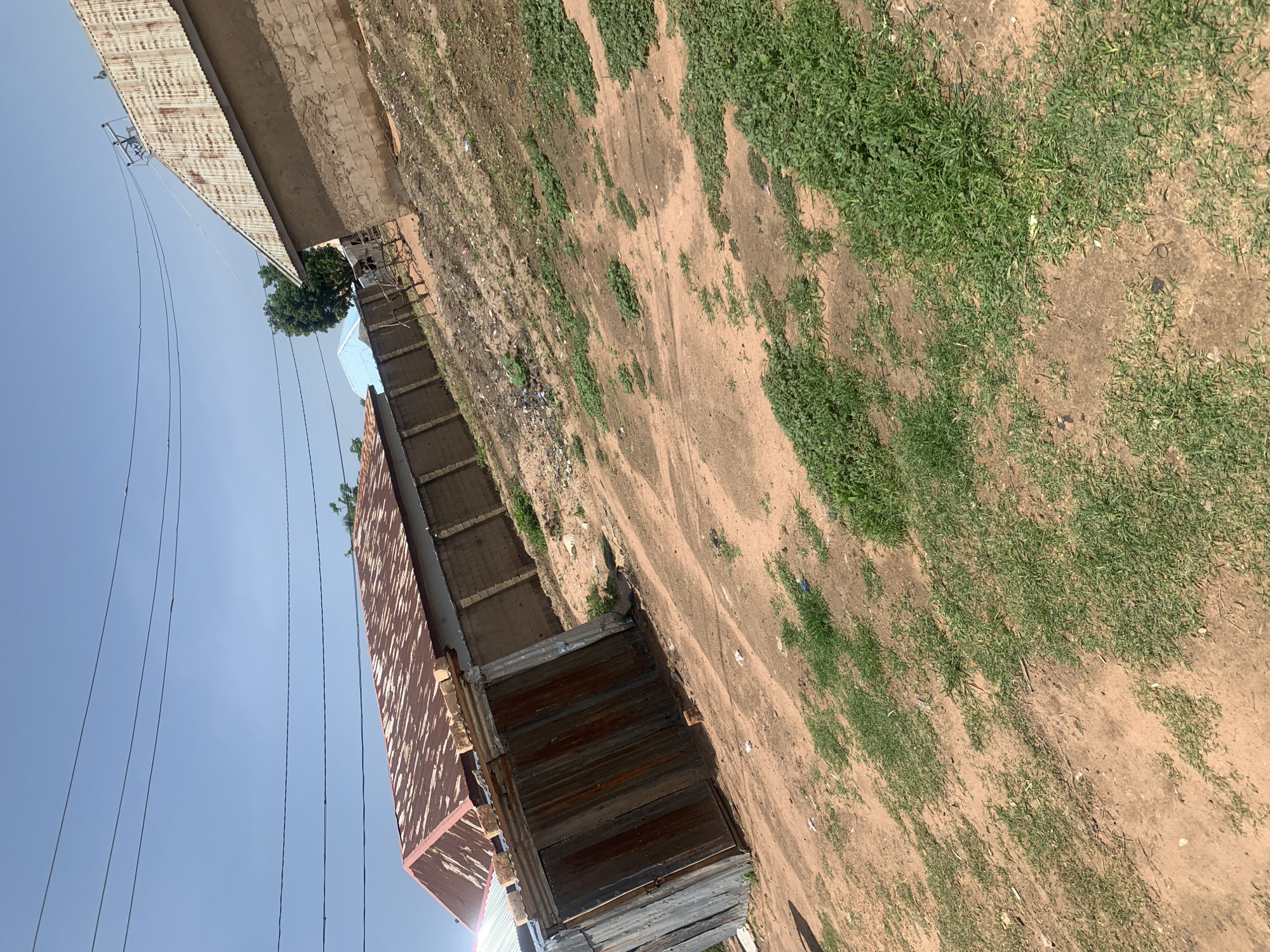 The image depicts an outdoor scene in Jauro Yinu, Nigeria, characterized by a dusty, uneven ground with patches of green grass. Several structures are visible, including buildings with corrugated metal roofs that appear weathered and rusted, and others constructed with brick or concrete blocks. A small, rudimentary shelter made of corrugated metal sheets is positioned in the foreground. Electrical wires are strung across the clear blue sky, suspended from poles. The overall environment suggests a developing or rural area with modest infrastructure. No people or specific activities are evident in the image.