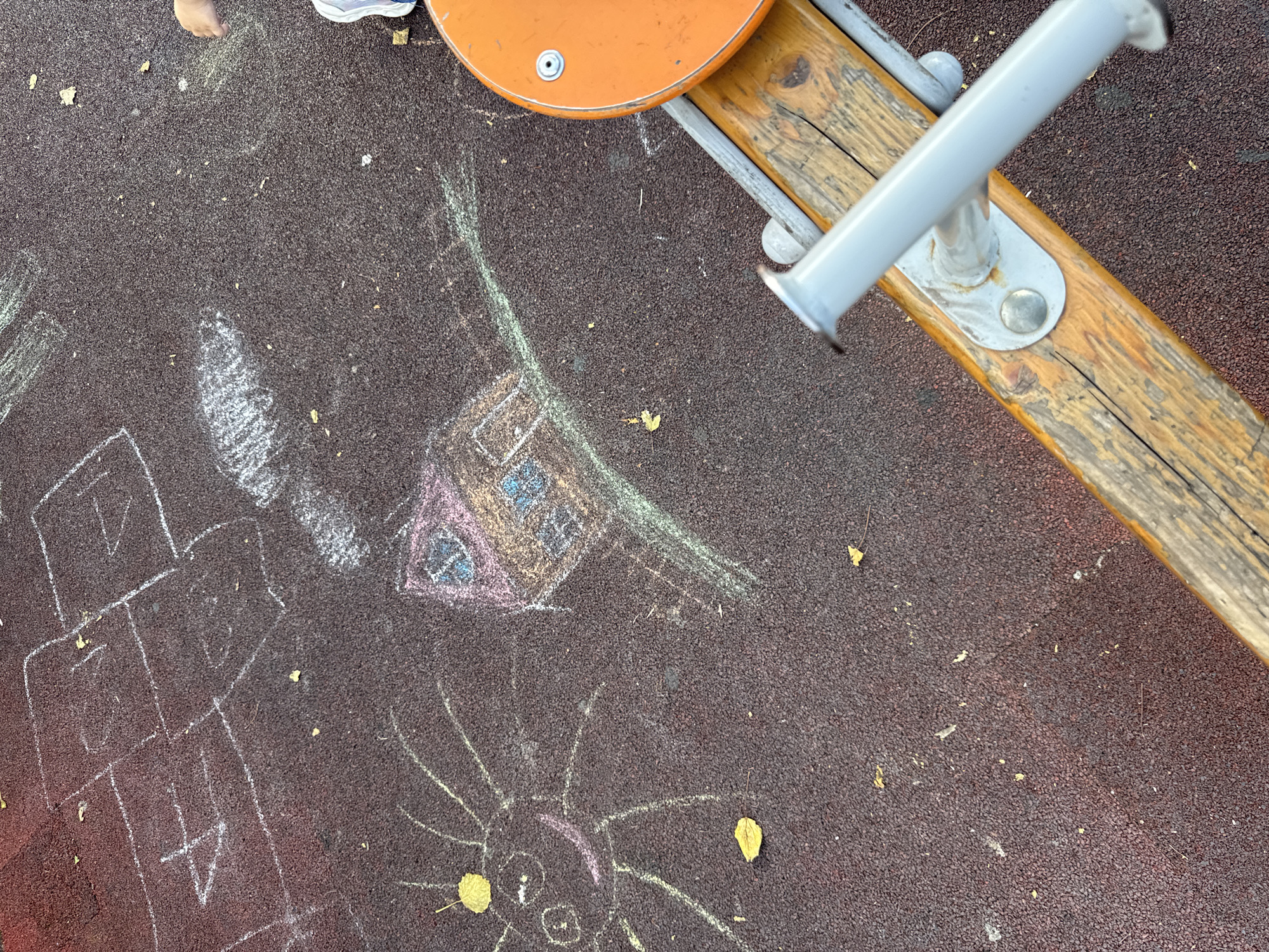 A child's drawing of a house and a sun is chalked on a dark reddish-brown rubberized playground surface. The drawing is located in the city of Vyazniki, Russia. A portion of an orange seesaw seat and its wooden and metal support structure are visible in the lower right quadrant of the image. The drawing of the house has a pink roof and blue windows. The sun is yellow with rays extending outwards. Several fallen leaves are scattered on the ground. A child's hand and shoe are partially visible in the upper right corner.