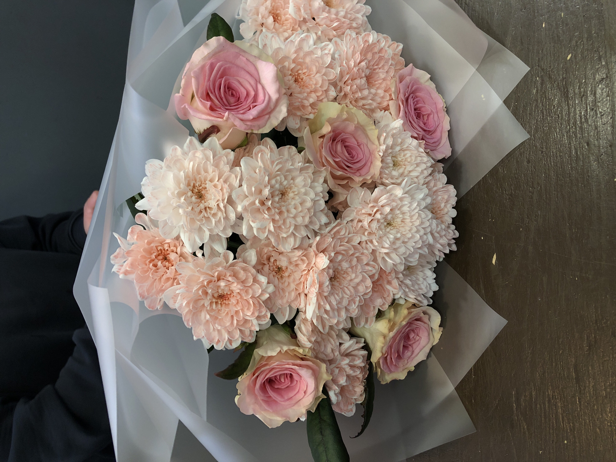 The image shows a bouquet of pink roses and chrysanthemums wrapped in white translucent paper. The bouquet is composed of numerous flowers, with the roses displaying a soft pink hue and the chrysanthemums exhibiting a blush or peach tone. The flowers are arranged densely, creating a full and rounded shape. The wrapping paper is folded and creased, giving the bouquet a structured appearance. The background includes a grey wall and a dark brown, possibly wooden, surface. There is no indication of the city of Chaikovsky or any specific location.