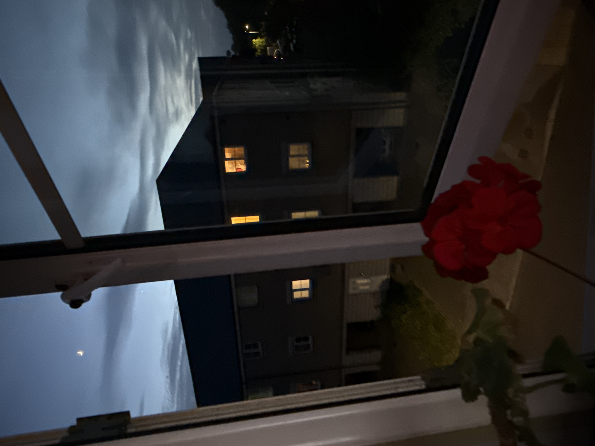 A nighttime scene is viewed through a window with a red flower in the lower right corner. The window reflects an apartment building with illuminated windows across the street, the clouds are visible through the sky reflected as well, and the moon can be seen near the bottom left corner of the image. The specific location within Great Yarmouth, United Kingdom is unable to be deduced.