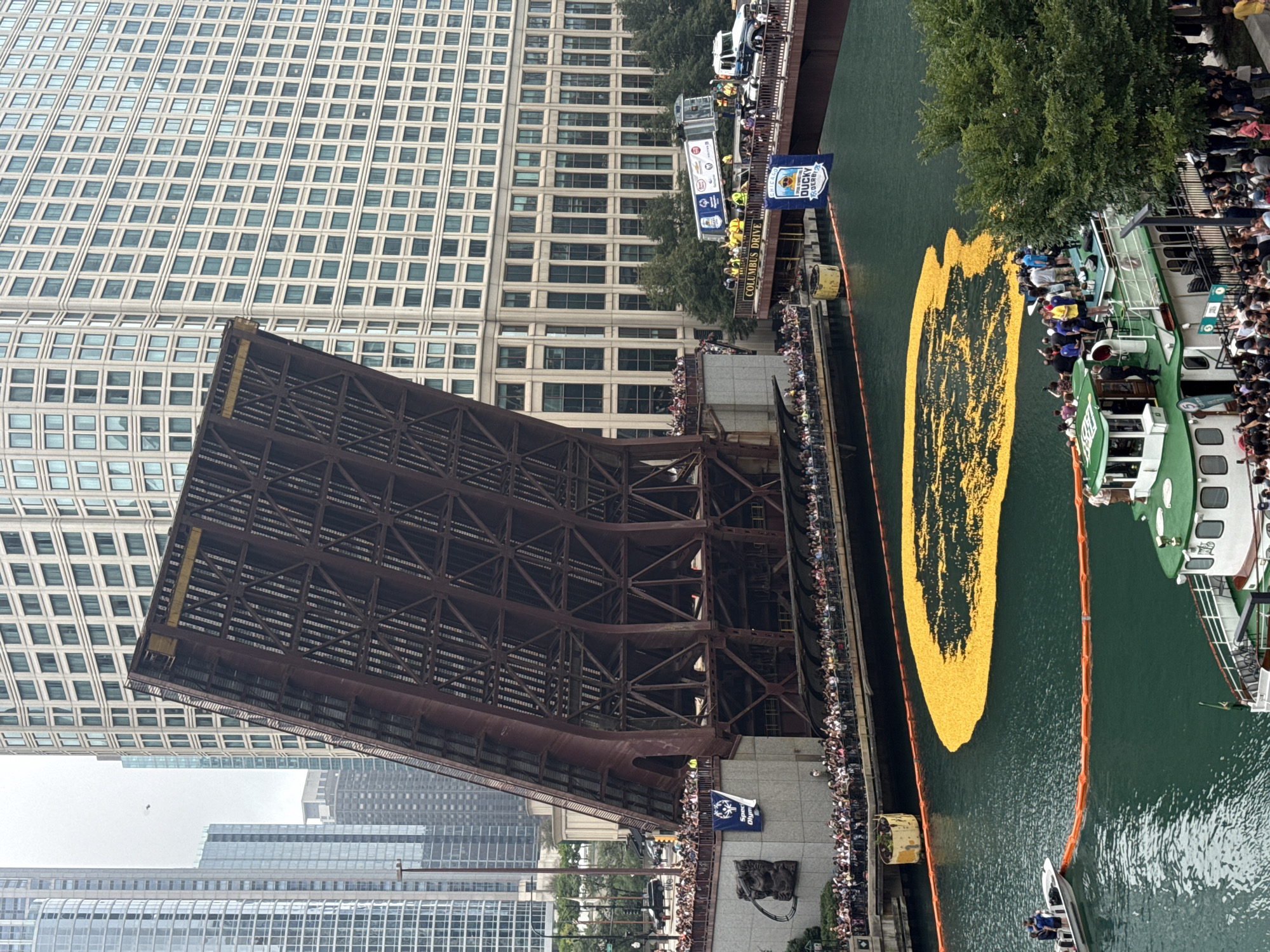 The image shows a drawbridge raised over the Chicago River in Chicago, Illinois, USA. Thousands of yellow rubber ducks are floating in the river, forming a large cluster. A banner proclaiming "Chicago Ducky Derby" hangs from a bridge in the background. Crowds of people are gathered on bridges and along the riverbanks to watch the event. Several boats are on the river, including a tour boat filled with people and a smaller boat with a few individuals. The scene is framed by modern high-rise buildings.