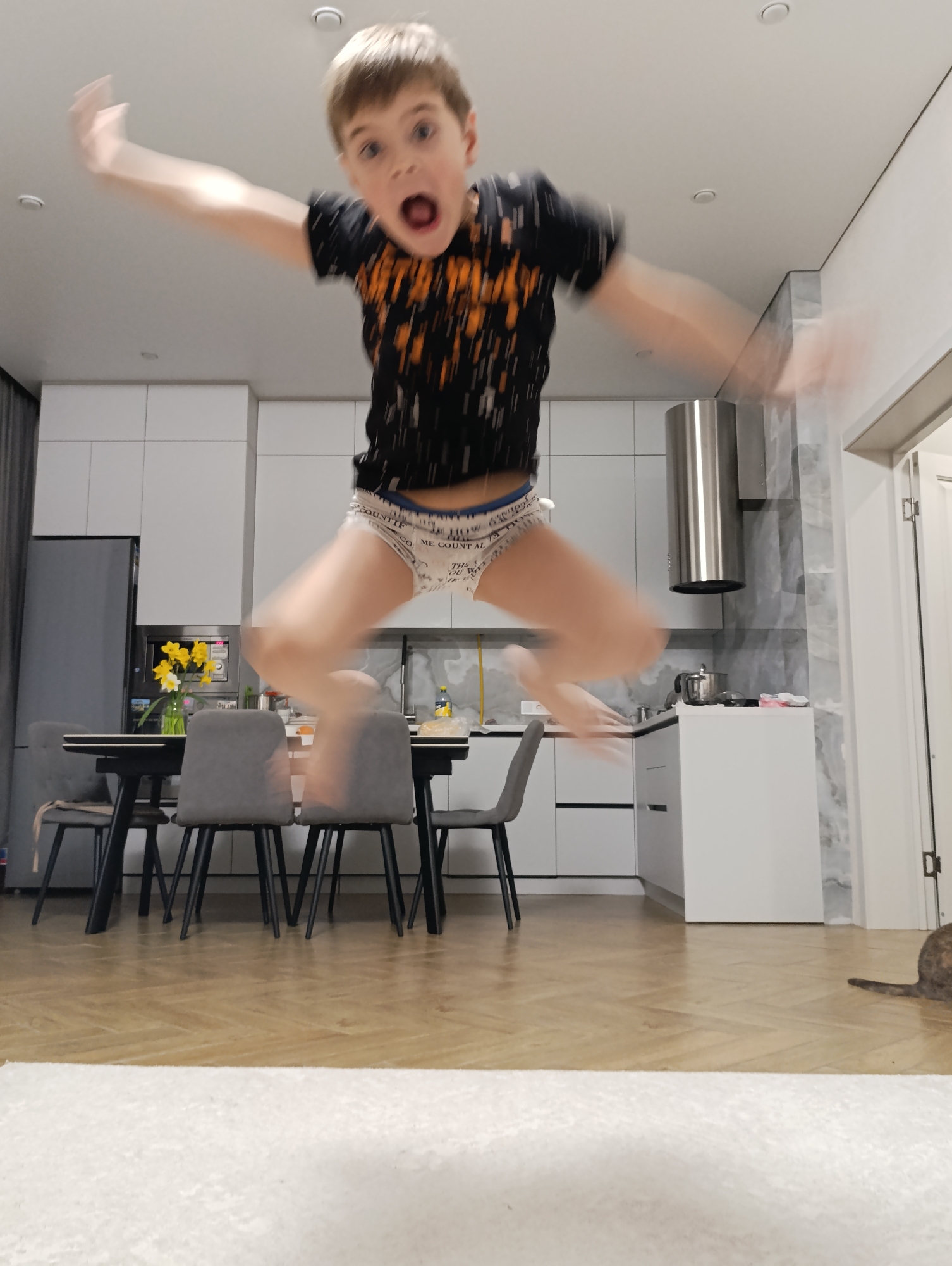 A child is jumping in a kitchen. The kitchen has white cabinets, a stainless steel range hood, and a gray marble backsplash. There is a table with gray chairs and yellow flowers on it. The floor is wood and covered with a white rug. A cat is lying on the floor near the doorway. The location of the image is presumed to be Chaikovsky, Russia.