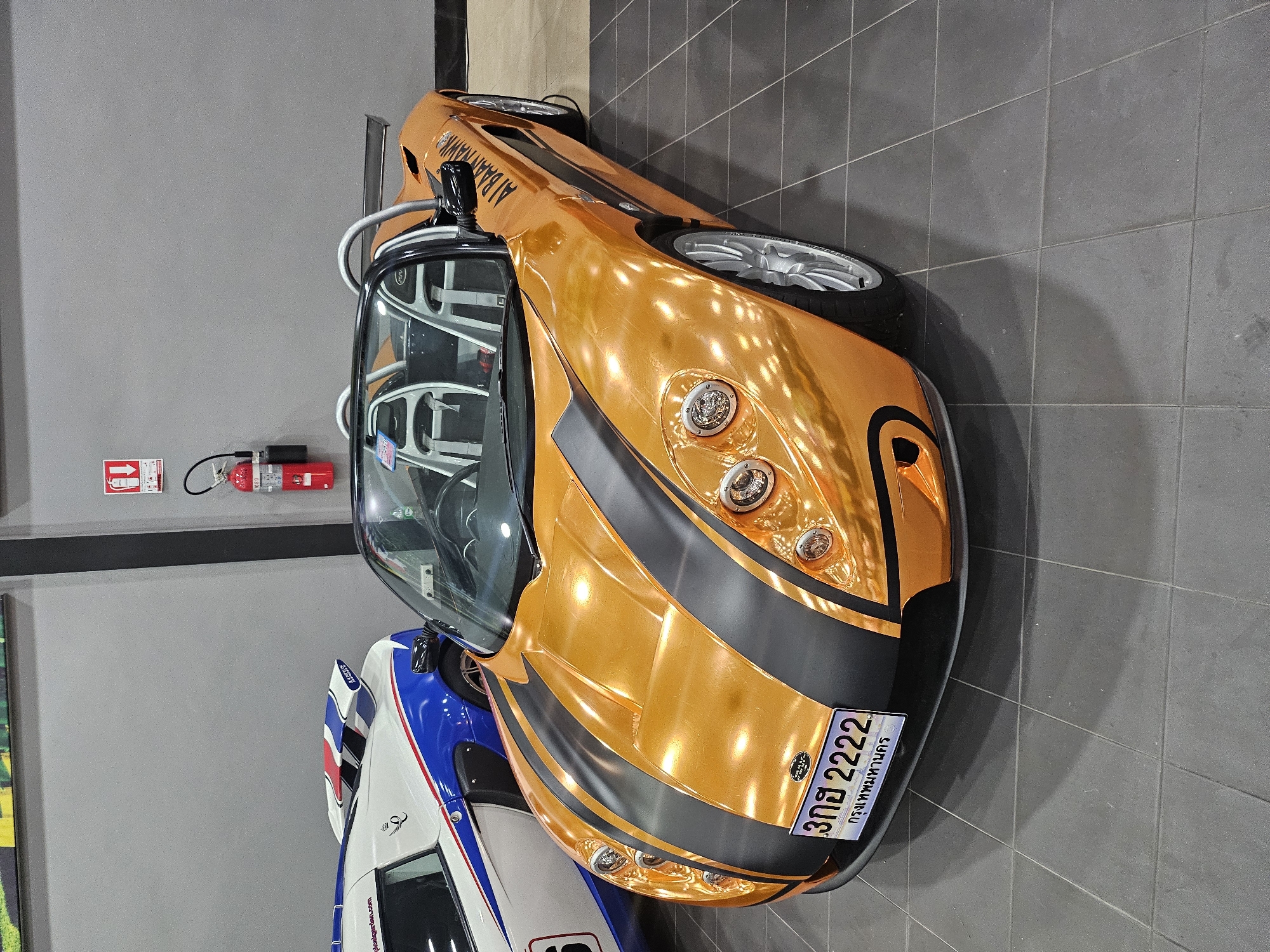A bright orange convertible sports car with black racing stripes is parked on a dark tiled floor in what appears to be a showroom or garage. The car has a license plate that reads "3กฮ 2222" and "กรุงเทพมหานคร" (Bangkok). A white and blue sports car with a red, white, and blue spoiler is parked to the left of the orange car. In the background, a red fire extinguisher hangs on a grey wall. The scene is indoors, with overhead lighting reflecting off the glossy finish of the orange car. The overall impression is one of automotive display.