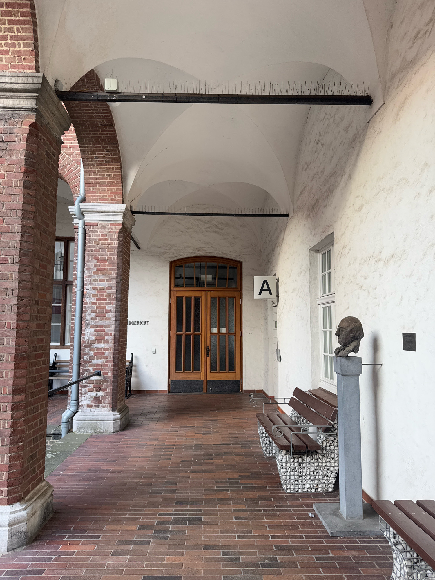 The image shows the arched walkway of a building with red brick pillars and a white, textured wall. A large wooden double door is visible in the background. To the left of the door, text on the wall reads "DGERICHT". A white sign with a black letter "A" hangs on the wall to the left of the door. There is a bust on a pedestal to the right of the door, with two benches in front of it. The walkway is paved with red brick. The scene appears to be in Cleves, Germany, specifically at the Amtsgericht (District Court).