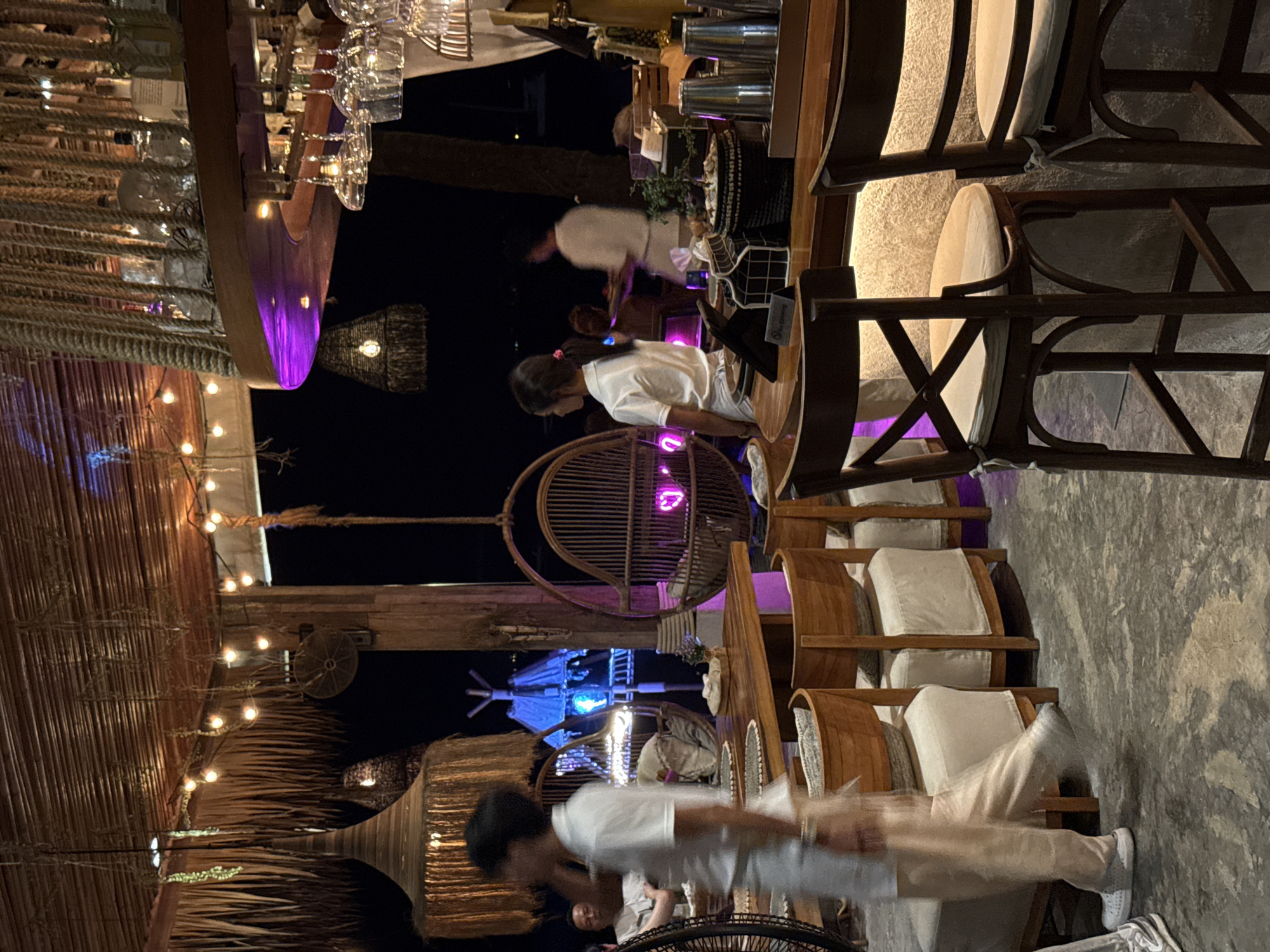 This is a medium shot, taken at night, of a dimly lit, open-air restaurant or bar area. The ceiling is covered with a woven thatch material, and hanging from it are rustic lamps and what appear to be decorative dried foliage. String lights are draped across the ceiling, providing a warm, ambient glow.
In the foreground, a person in a white polo shirt and light-colored pants is walking, motion blurred, from left to right. They are on a grey, textured floor. To the right of this person, a wooden table is set with placemats and what appears to be food. Behind the table are several light-colored upholstered seating areas, some with wooden chairs.
Further into the background, a person in a white uniform is standing near a bar area. The bar has a curved wooden countertop and is lined with bar stools. Behind the bar, bottles and glassware are visible. There are also some metallic containers and a small potted plant on the bar.
To the left and center background, there are decorative hanging wicker chairs with cushions, one of which has a neon pink heart shape displayed within it. In the far background, there is a glimpse of a structure with blue lights, possibly a tent or another building. The overall atmosphere is tropical and relaxed, with a focus on natural materials and ambient lighting.
The scene is located in Baan Bo Phut, Thailand, suggested by the tropical aesthetic and likely the style of the establishment.