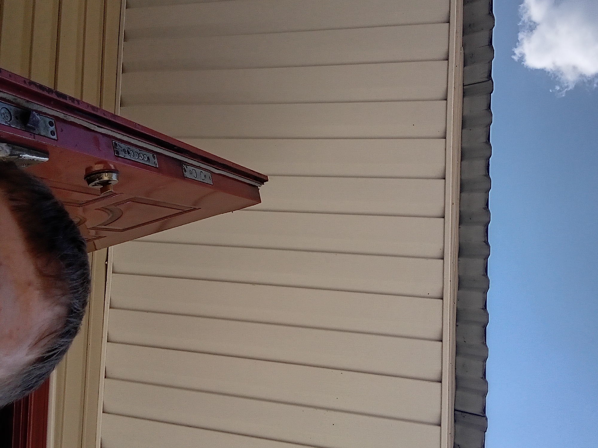 The image shows a close-up, low-angle view of a partially open red door, with the top of a person's head visible in the foreground. The door appears to be made of wood and has visible hardware, including what appear to be lock mechanisms and a round doorknob. The background consists of a light-colored, vertically slatted structure, a corrugated metal roof, and a clear blue sky with a single white cloud. This scene was captured in Verkhniachka, Ukraine.