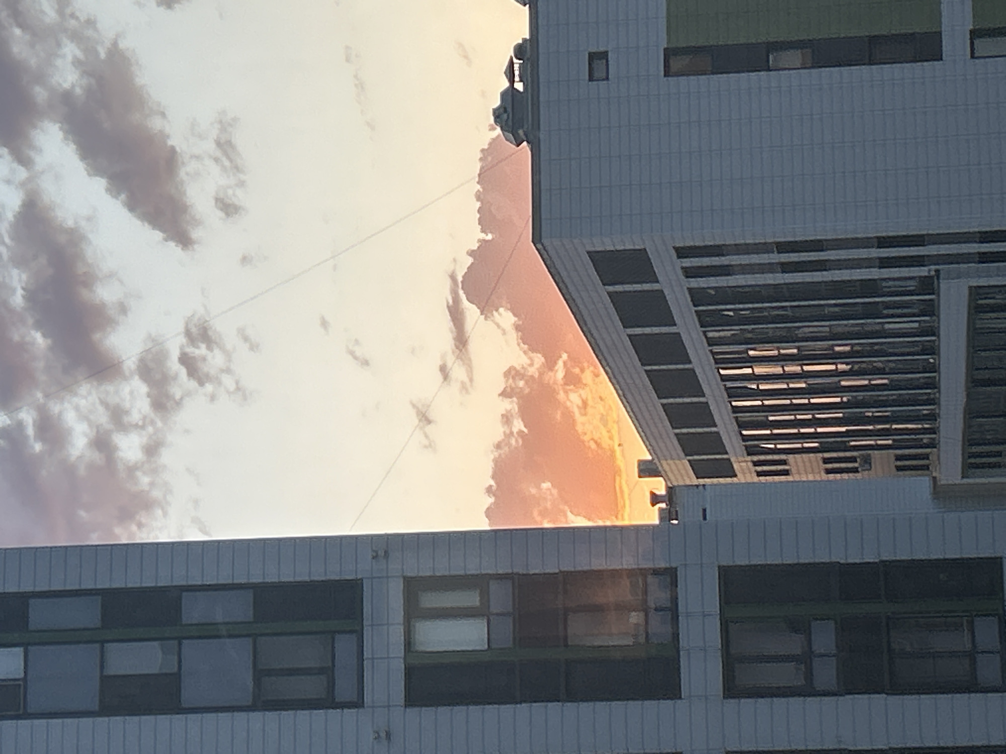 The image depicts a cityscape at sunset, with the sky painted in hues of orange and pink from the setting sun illuminating the clouds. Two modern buildings are prominently featured. The building on the left is tall and slender, with a facade of light-colored panels and dark-tinted rectangular windows arranged vertically. The building on the right is wider and lower, also constructed with light-colored panels, featuring rows of dark windows that reflect the warm light of the sunset. The top of this building has visible ventilation shafts and a small rectangular window. Fine wires are visible stretching across the sky. The scene is urban and contemporary, suggestive of a developed city. Based on the architectural style and urban setting, this image is likely from Astana, Kazakhstan.