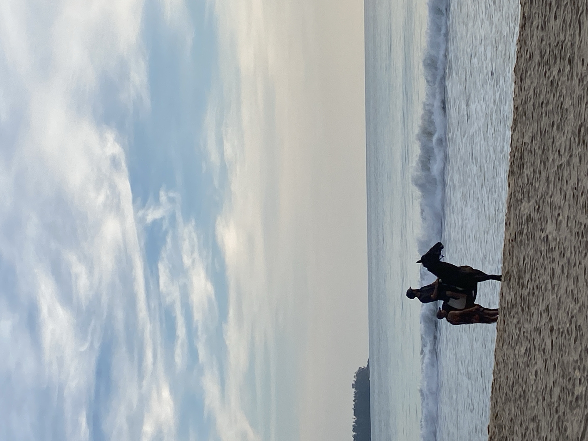 A daytime, landscape-oriented photograph shows a beach scene with a cloudy sky dominating the left half of the frame. A body of water, presumably the ocean, extends from the top to the bottom of the frame. Near the bottom right corner of the image, there is a black horse being ridden by a man. A second man stands close to the horse. The beach area occupies the rightmost portion of the image. The location is believed to be Choeng Thale, Thailand.