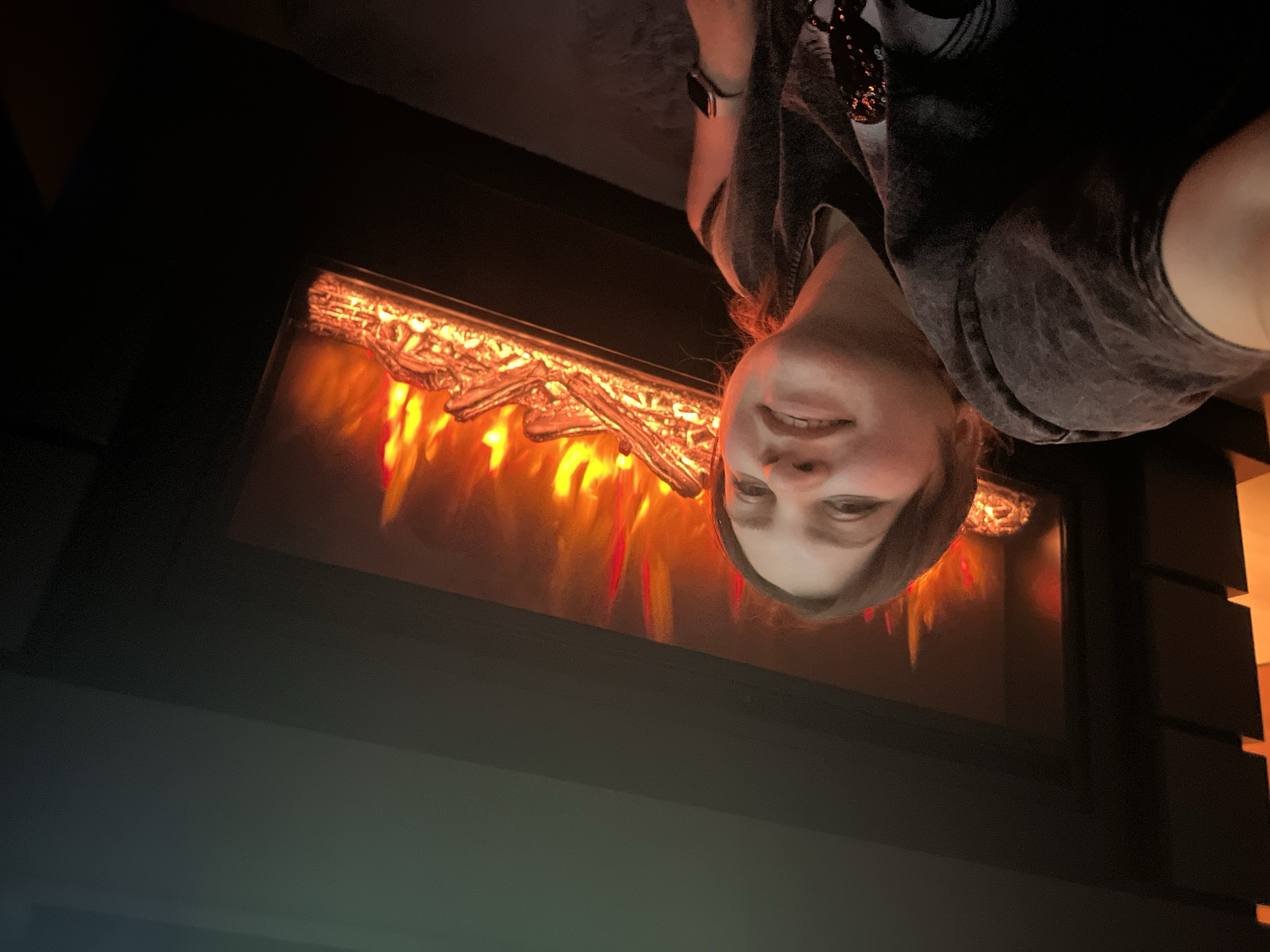 A woman wearing a gray t-shirt is smiling in front of an electric fireplace with a faux log set and glowing red "flames". The image appears to be taken indoors, possibly in Perm, Russia. The scene is dimly lit, with the fireplace providing the main source of illumination.