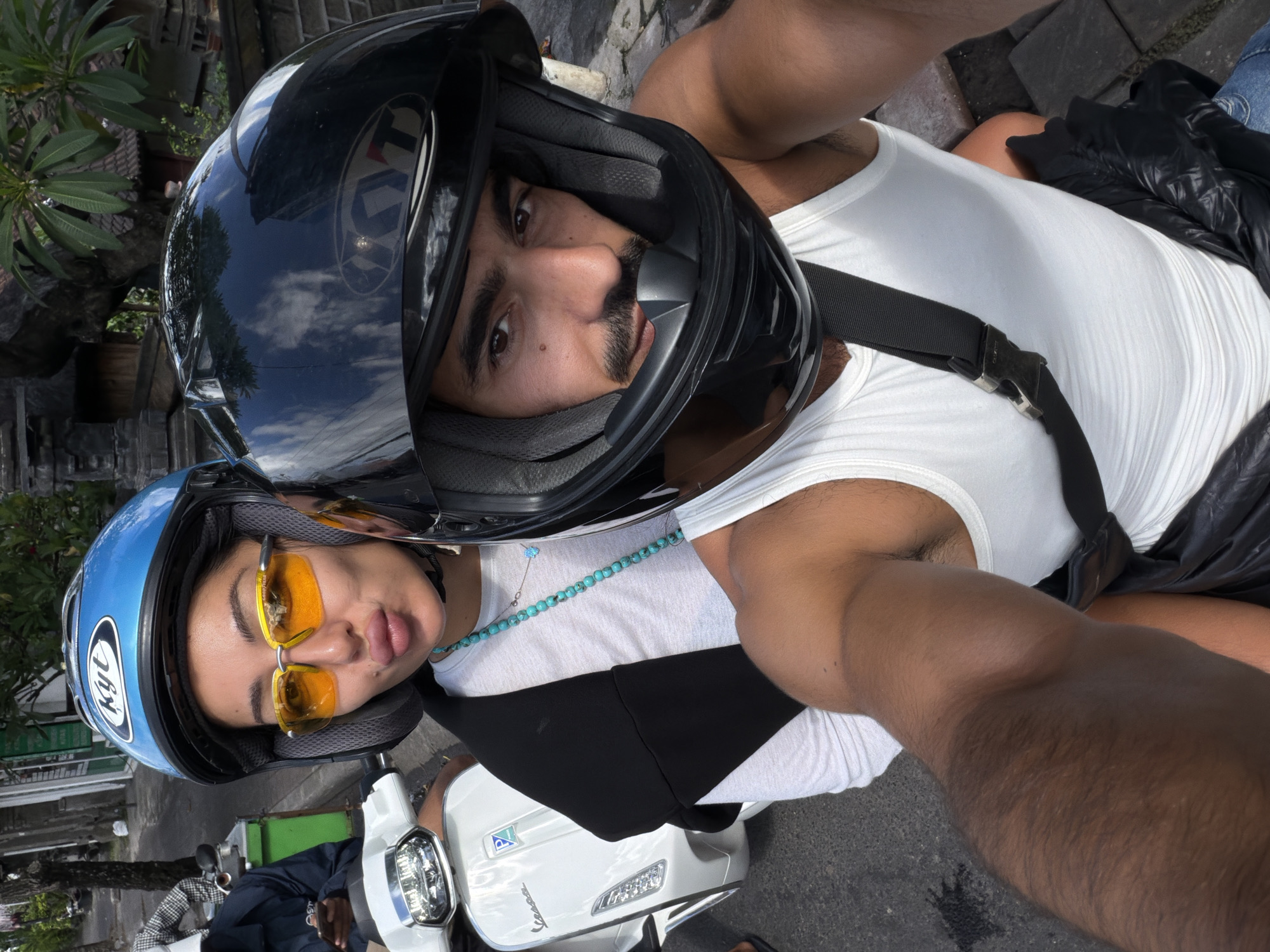 Two individuals wearing helmets are sitting on a white Vespa scooter. The person in the front is wearing a black helmet and a white tank top, with a black strap crossing their chest. The person behind is wearing a blue helmet and yellow sunglasses, also with a white shirt and turquoise necklace. The background shows a street scene, with buildings and foliage visible. The location is North Kuta, Indonesia.