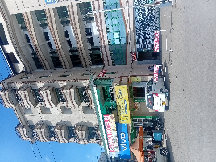 The image shows a street scene in front of a large, multi-story building in Mlolongo, Kenya, located at coordinates -1.394 latitude and 36.94 longitude. The building appears to be a mix of commercial and residential use, with businesses on the lower levels and apartments above. The ground floor is occupied by several shops, including a Vivo mobile phone store and a restaurant called "MASH Pee". There is a white car parked in front of the restaurant, and a red sign that reads "AUTHORIZED VEHICLES ONLY" is visible. There are several people in the scene, including a man sitting on a bench in front of the building, a group of people standing in front of the Vivo store, and a person standing in front of the parked car. The street is paved with interlocking bricks and there is an orange awning tent visible in the lower left corner of the image. The building is made of concrete and has a beige exterior with brown window frames. The upper floors have balconies with white railings. The sky is blue and clear. A second red sign that reads "AUTHORIZED VEHICLES ONLY, NO PARKING" is in the foreground of the image.