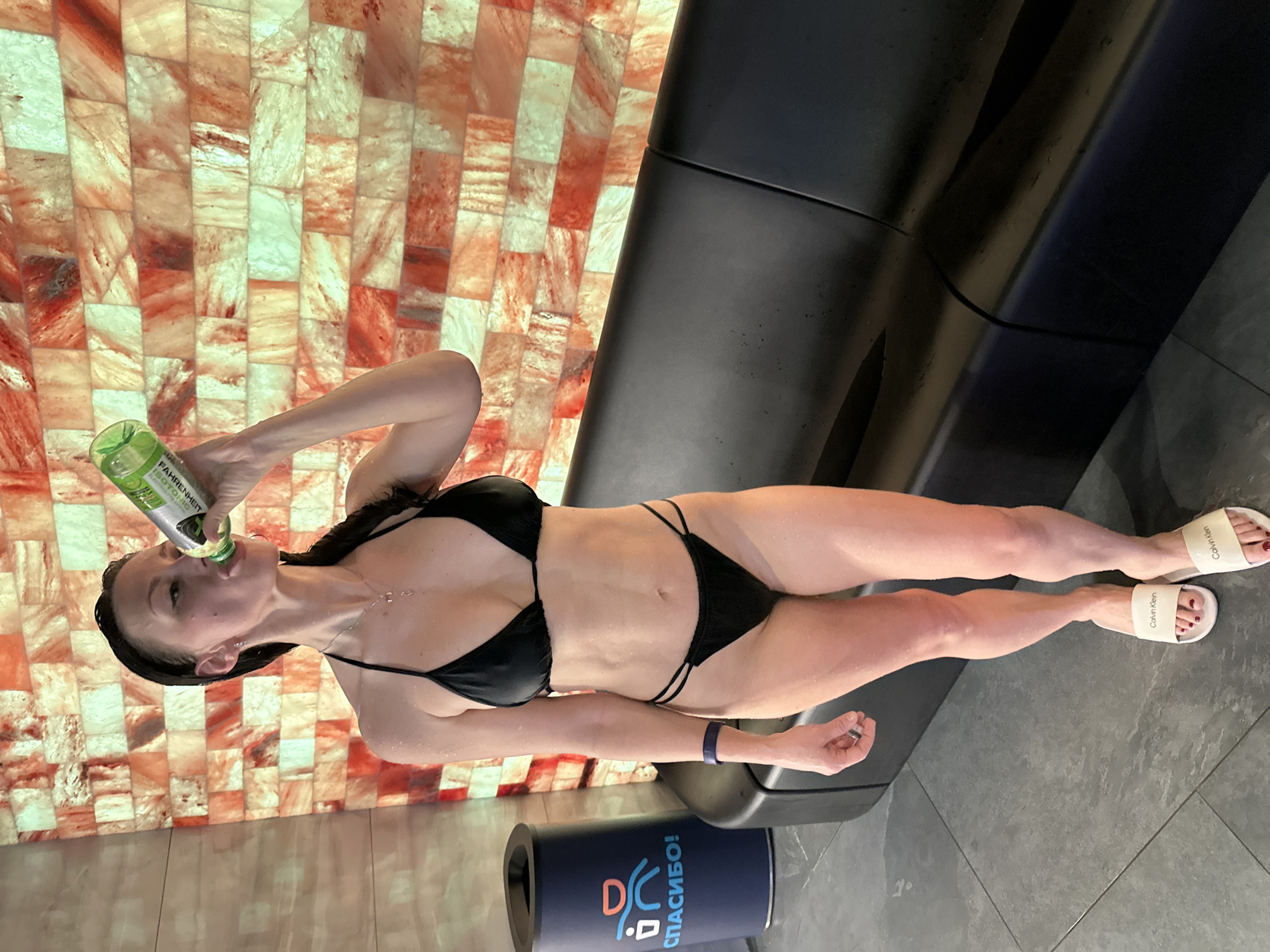 A woman in a black bikini is drinking from a green bottle while lying on a dark, modern bench in a room with a wall of illuminated salt bricks. She is wearing white Calvin Klein slide sandals and a fitness tracker on her left wrist. A dark blue cylindrical container with the word "CПАСИБО!" (Thank You!) and stylized figures is at the edge of the bench. The scene is inside a salt room at the Premier Sport fitness club in Saint Petersburg, Russia.