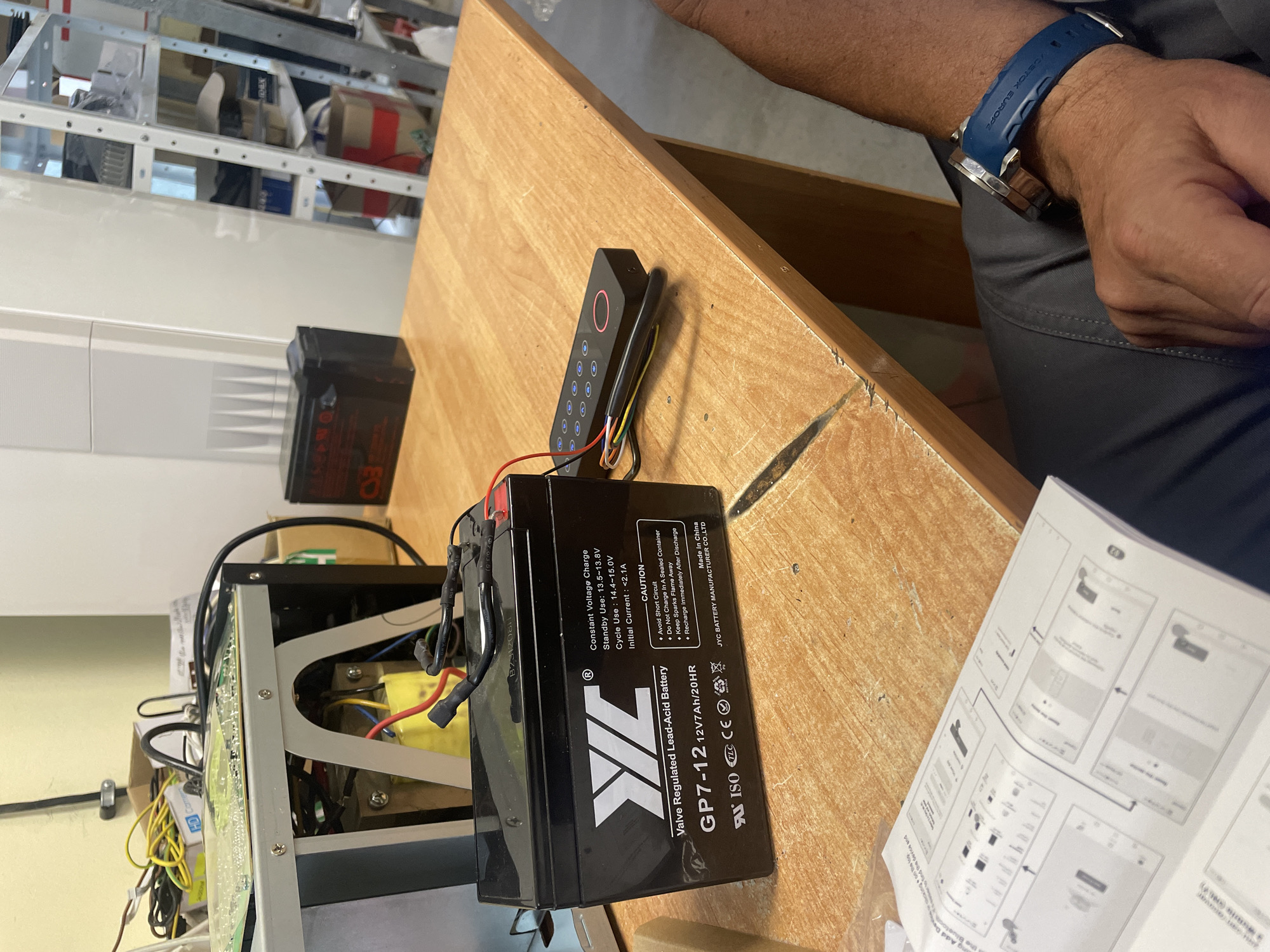 The image shows a close-up, top-down view of an electronics assembly or setup taking place on a wooden table. A person's arm, wearing a blue watch with "DETOX EUROPE" visible on the strap, and their leg in grey pants are visible on the right side of the frame, suggesting they are actively involved in the work.
At the center of the foreground is a black, rectangular "XL" brand "Valve Regulated Lead-Acid Battery," model "GP7-12," clearly labeled with specifications: "12V 7Ah/20HR." Further details on the battery include "Constant Voltage Charge," "Standby Use: 13.5~13.8V," "Cycle Use: 14.4~15.0V," and "Initial Current: <2.1A." It also provides "CAUTION" notes such as "Avoid Short Circuit," "Do Not Charge In A Sealed Container," "Keep Sparks Flame Away," and "Recharge Immediately After Discharge." The battery is "Made in China" by "JYC BATTERY MANUFACTURER CO.,LTD" and carries ISO, CE, and TLC certifications.
Adjacent to the battery is a compact black electronic device, possibly a keypad or access control unit, featuring an array of circular buttons (likely numbered 0-9) and a prominent red or pink illuminated ring at its top. Wires of various colors (red, yellow, green, orange, blue, white) are connected to this device. To the left of the battery, the open casing of another electronic unit or power supply reveals internal components, including transformers and circuitry.
An open instruction manual or guide is spread out on the table to the right, displaying diagrams and text related to "Add Device" and "Bluetooth," with the page number "20" visible. The wooden table surface exhibits signs of wear and minor cracks. The background shows metal shelving units stocked with various items, indicating a workshop or technical storage area. The scene depicts a detailed process of setting up or troubleshooting electronic equipment.