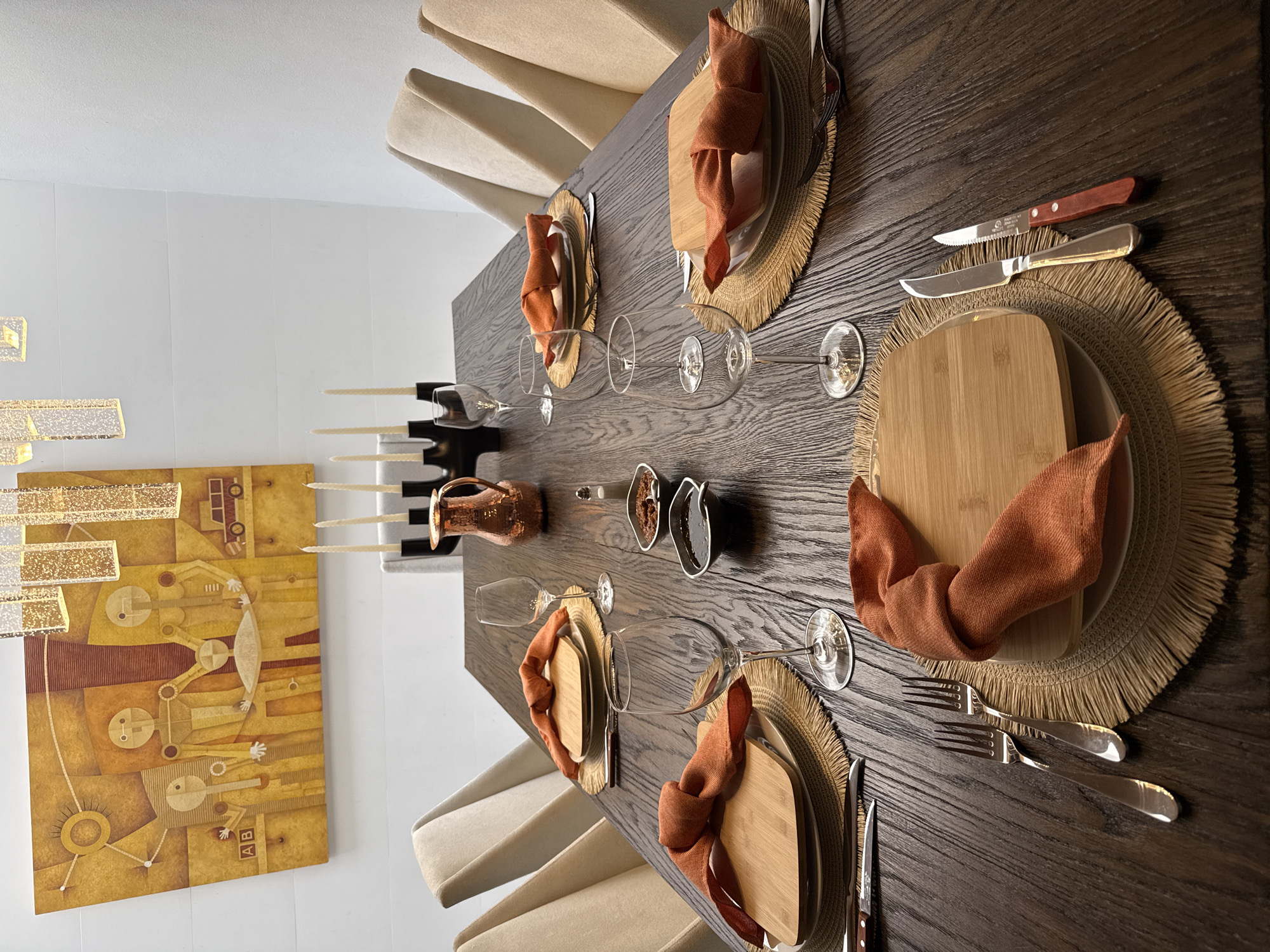 This media file presents a contemporary dining room setup, likely within a home in Huixquilucan de Degollado, Mexico. The central focus is a large, dark brown wooden dining table with a visible grain, meticulously set for a meal.
Around the table are six modern dining chairs, upholstered in a light beige fabric. Each place setting features a round, woven straw placemat with frayed edges, a light-colored dinner plate, and a rectangular bamboo serving board. Burnt orange linen napkins are artfully folded on top of the boards. The cutlery at each setting includes a serrated steak knife with a wooden handle, a standard metallic knife, and two forks of different sizes. One steak knife bears the visible text "TRAMONTINA BRASIL INOKS STAINLESS STEEL." Two wine glasses, of differing shapes, are placed at each setting.
The table's centerpiece consists of a copper-toned pitcher, a modern black candelabra holding several unlit white taper candles, and two small, oval dishes containing dark condiments or sauces.
The surrounding walls are clean and white. On the left, a large rectangular abstract painting in shades of yellow, gold, and orange depicts stylized figures and geometric shapes. Above the painting, modern light fixtures with sparkling, rectangular elements provide artificial illumination, suggesting the table is prepared for an evening gathering or meal. The overall scene conveys an atmosphere of sophisticated comfort and anticipation for a social event.