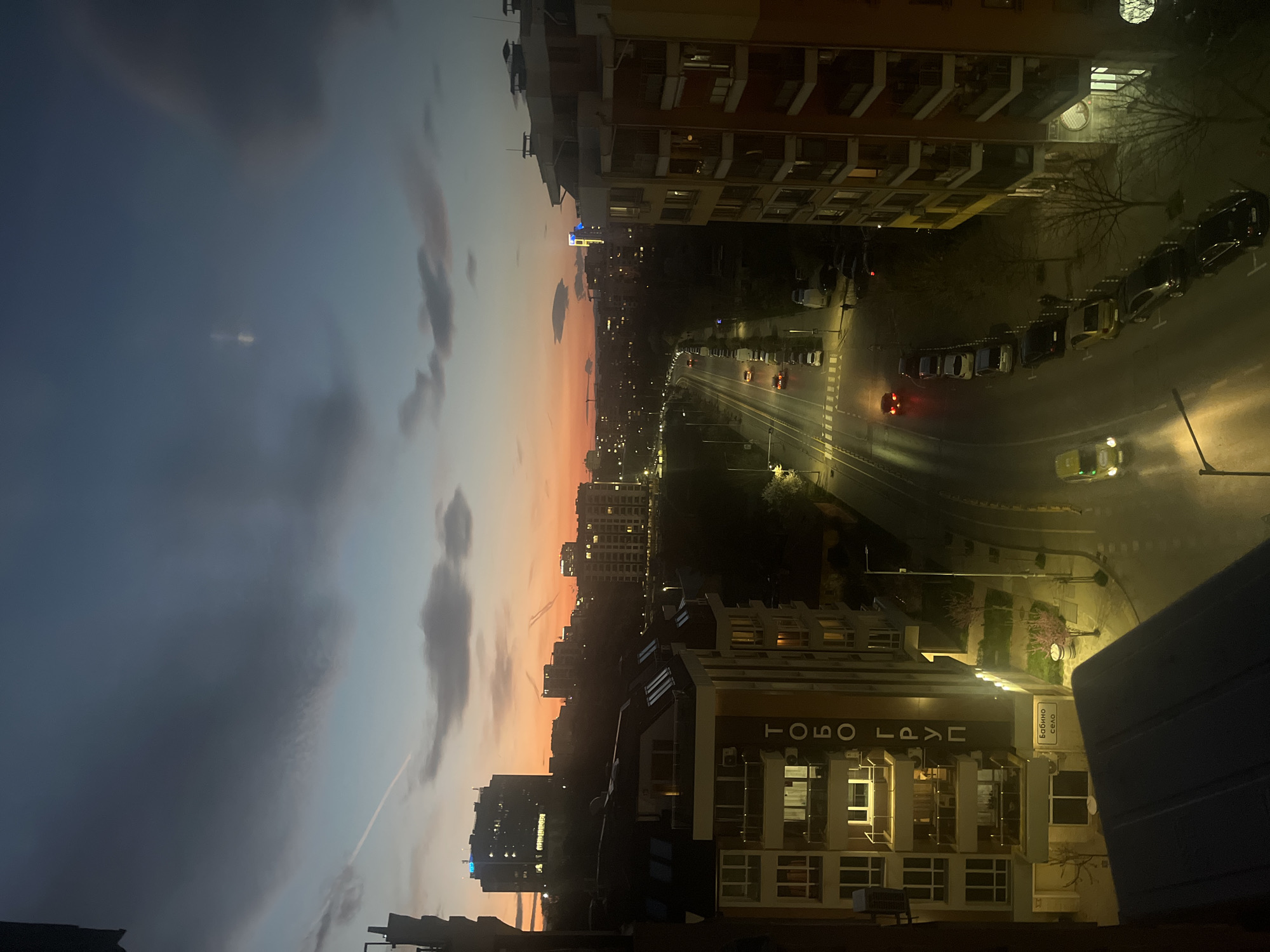 The image depicts a view of a city street at dusk, likely in Sofia, Bulgaria. Buildings are illuminated with warm lighting, and cars are driving on the street. The sky transitions from a dark blue to a warm orange, with clouds scattered throughout. The buildings on either side of the street are residential, with multiple floors and lit windows. The street has streetlights and marked lanes. A sign written in Cyrillic is visible on one of the buildings.