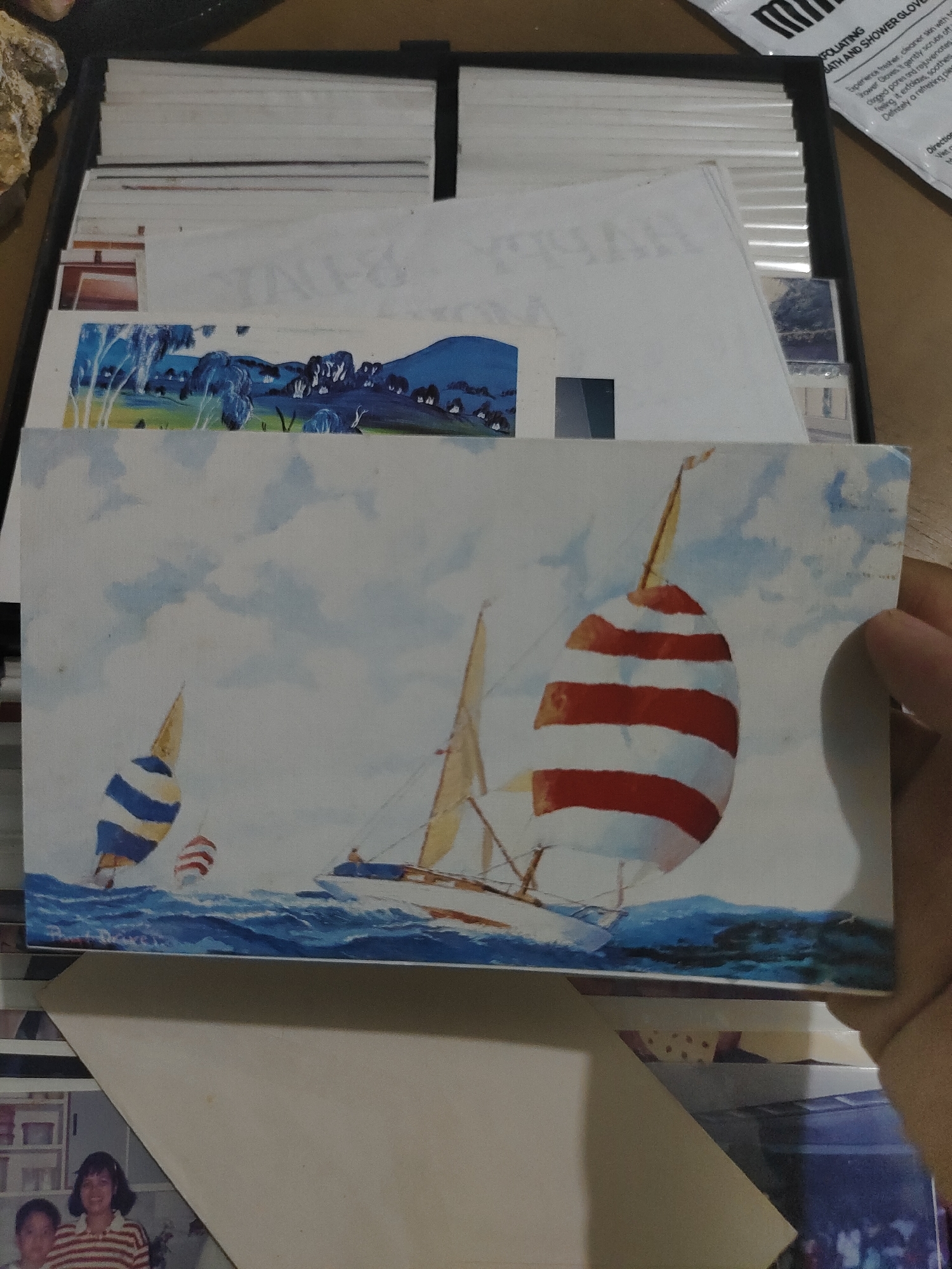 A forensic examination of the image reveals a human right hand holding a rectangular postcard depicting a painting of sailboats on water. The postcard features three sailboats: a prominent sailboat on the right with a red and white striped spinnaker, a smaller sailboat on the left with a blue and yellow striped spinnaker, and a distant smaller sailboat with a red and white striped sail. The water is rendered in shades of blue with white accents, indicating waves, under a light blue sky with white clouds.
Behind and partially obscured by the held postcard, a dark-colored container, possibly a photo box, is filled with various other postcards or photographs. One visible item in the box is a landscape painting featuring blue trees, green grass, and blue mountains. In the lower left foreground, beneath the held postcard, a photograph shows two individuals; one is a woman with dark hair wearing a red and white striped shirt, smiling, next to a partially visible child.
In the upper right background, a white package is visible, partially obscured, with the legible text "EXFOLIATING BATH AND SHOWER GLOVE." The upper left background includes a textured, light-brown object, possibly a rock or mineral specimen. The overall environment suggests an indoor setting, likely a table or desk where these items are laid out. The location is undetermined.