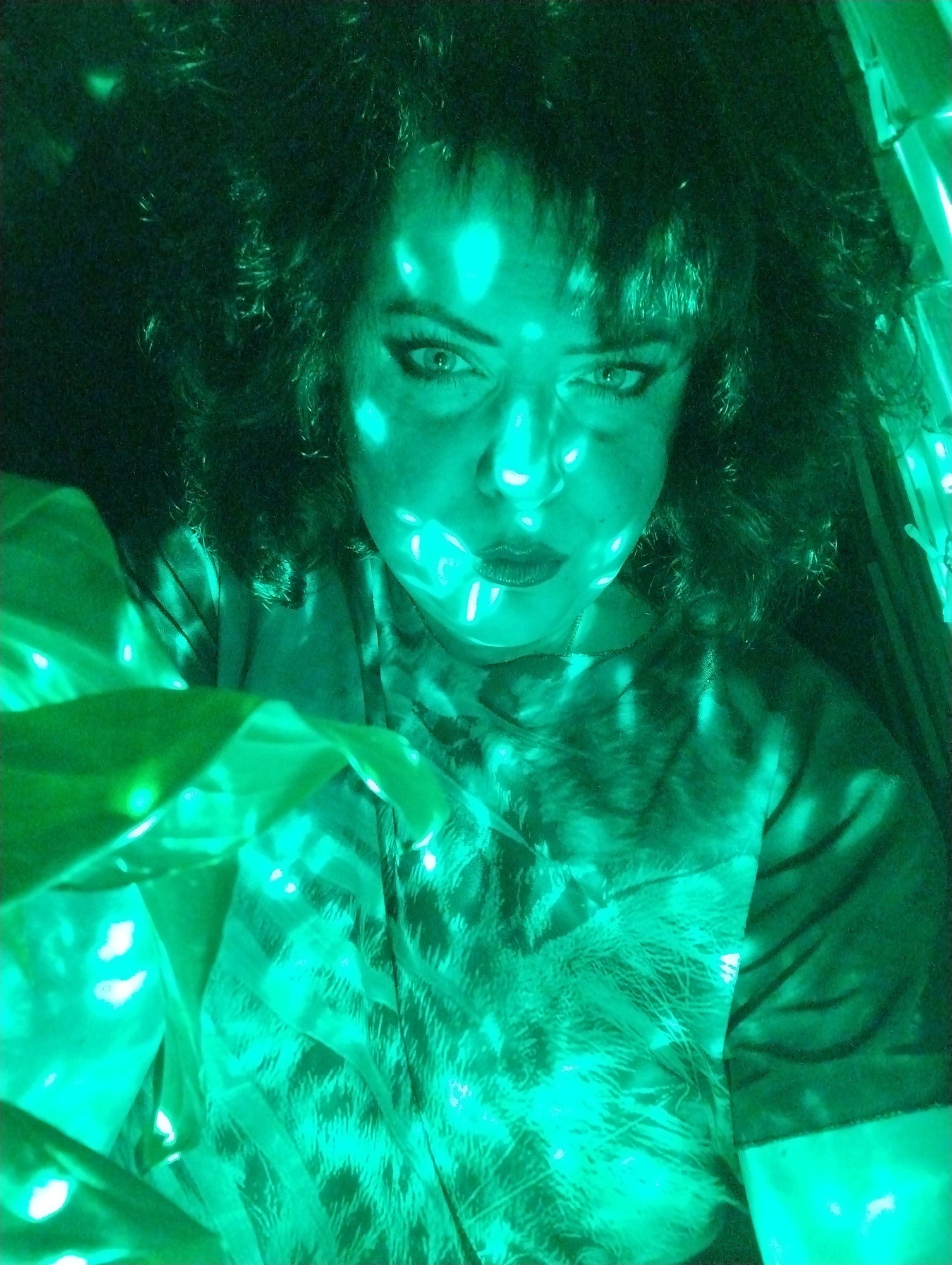 A woman is depicted in an indoor setting, bathed in green light. Her hair is dark and curly. She is wearing a shirt with a pattern reminiscent of an animal print. The light source creates a dappled effect on her face and clothing. The location is not evident from the image and cannot be determined to be Grishanki, Russia.
