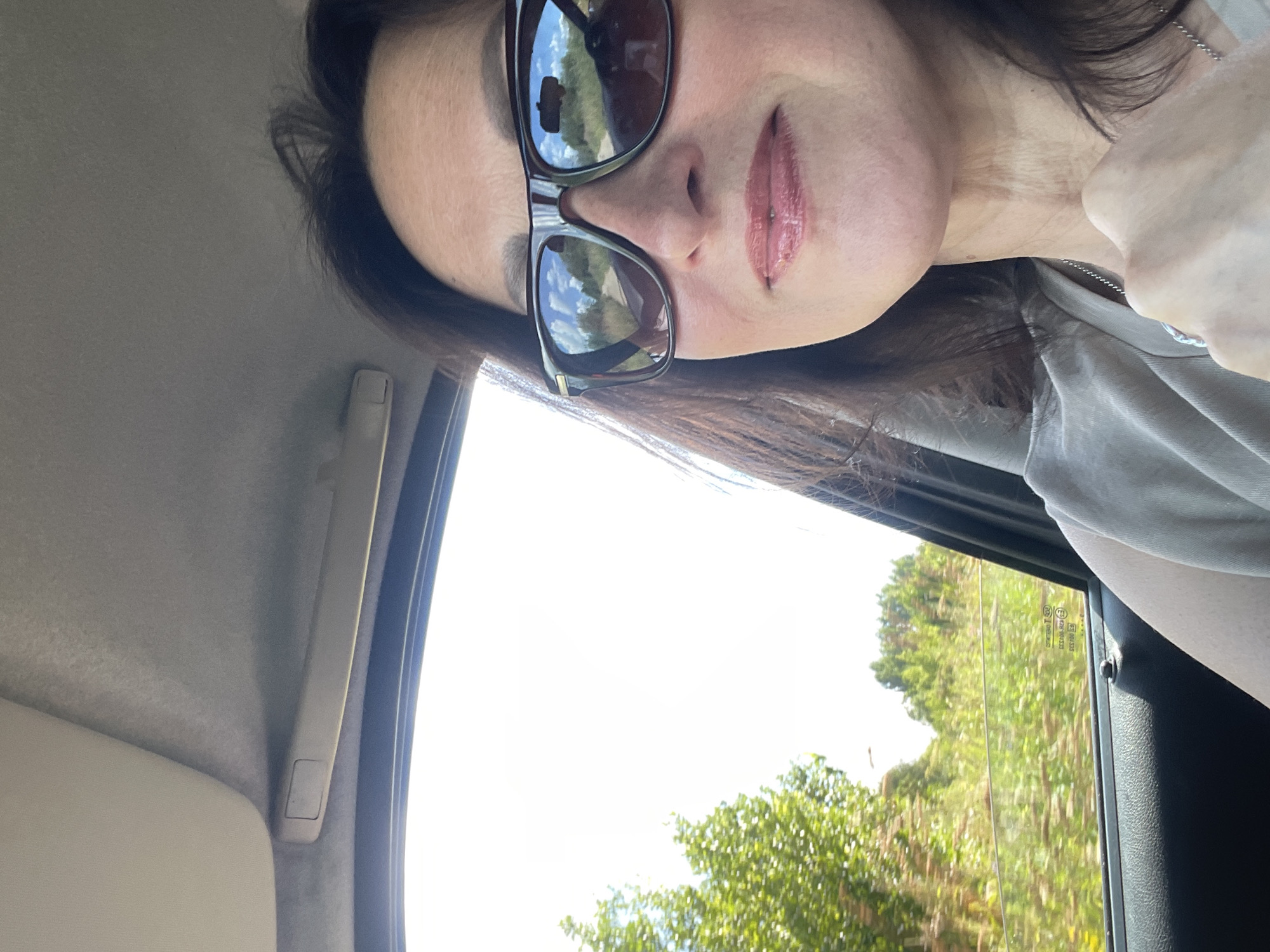 A medium shot photo shows a woman with dark hair and sunglasses, sitting in the passenger seat of a car. She is looking towards the camera, with her mouth slightly open and a hint of a smile. Her hair is dark brown and falls past her shoulders. She is wearing a light-colored t-shirt and a silver necklace. The sunglasses she is wearing have dark frames and are reflecting the sky and scenery outside the car. Through the car window, trees and greenery are visible. The interior of the car, including the headliner and door panel, are also visible. The image was taken in the city of Дресвяново, Russia.