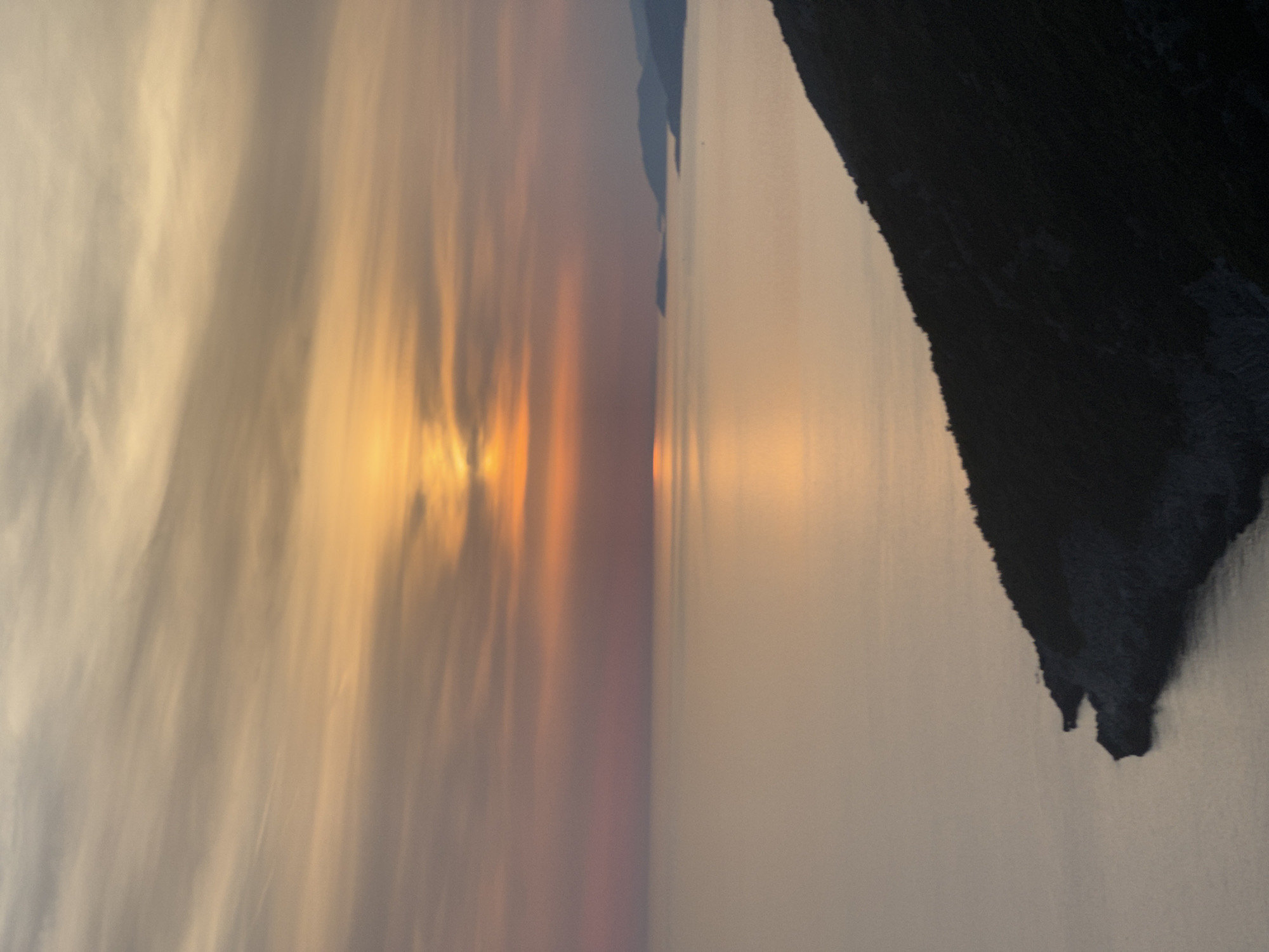 The image depicts a serene coastal landscape at sunset. The sky is ablaze with hues of orange, pink, and yellow, with soft, wispy clouds stretching across the horizon. A calm sea reflects the warm colors of the sky, creating a shimmering pathway of light. In the foreground, a dark, silhouetted hillside slopes down towards the water. In the distance, a chain of hazy, mountainous islands emerges from the sea, their forms softened by the fading light. The overall impression is one of tranquility and natural beauty, likely captured from an elevated vantage point overlooking the Adriatic Sea in the vicinity of Zagora, Montenegro.