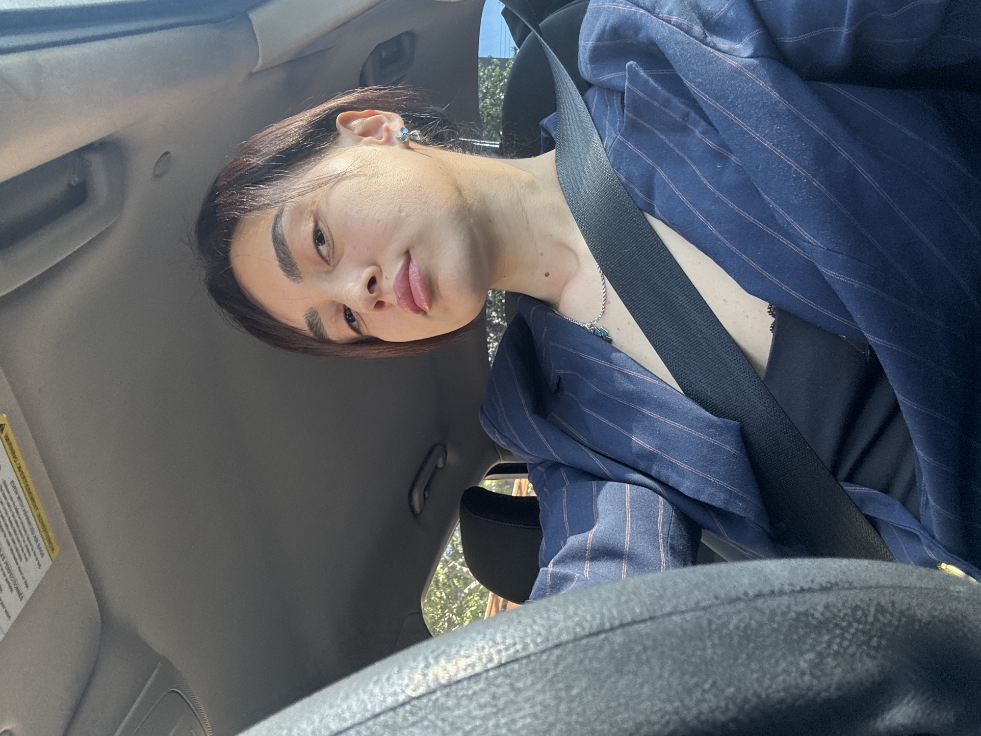 A woman wearing a navy pinstripe blazer and a black top is in a car, secured by a seatbelt. The photo is taken from a low angle, showing the steering wheel prominently. The woman has short brown hair and is wearing small earrings and a necklace. Sunlight filters through trees outside the car windows. The interior of the car is a light gray color. This image was captured in Tbilisi, Georgia.