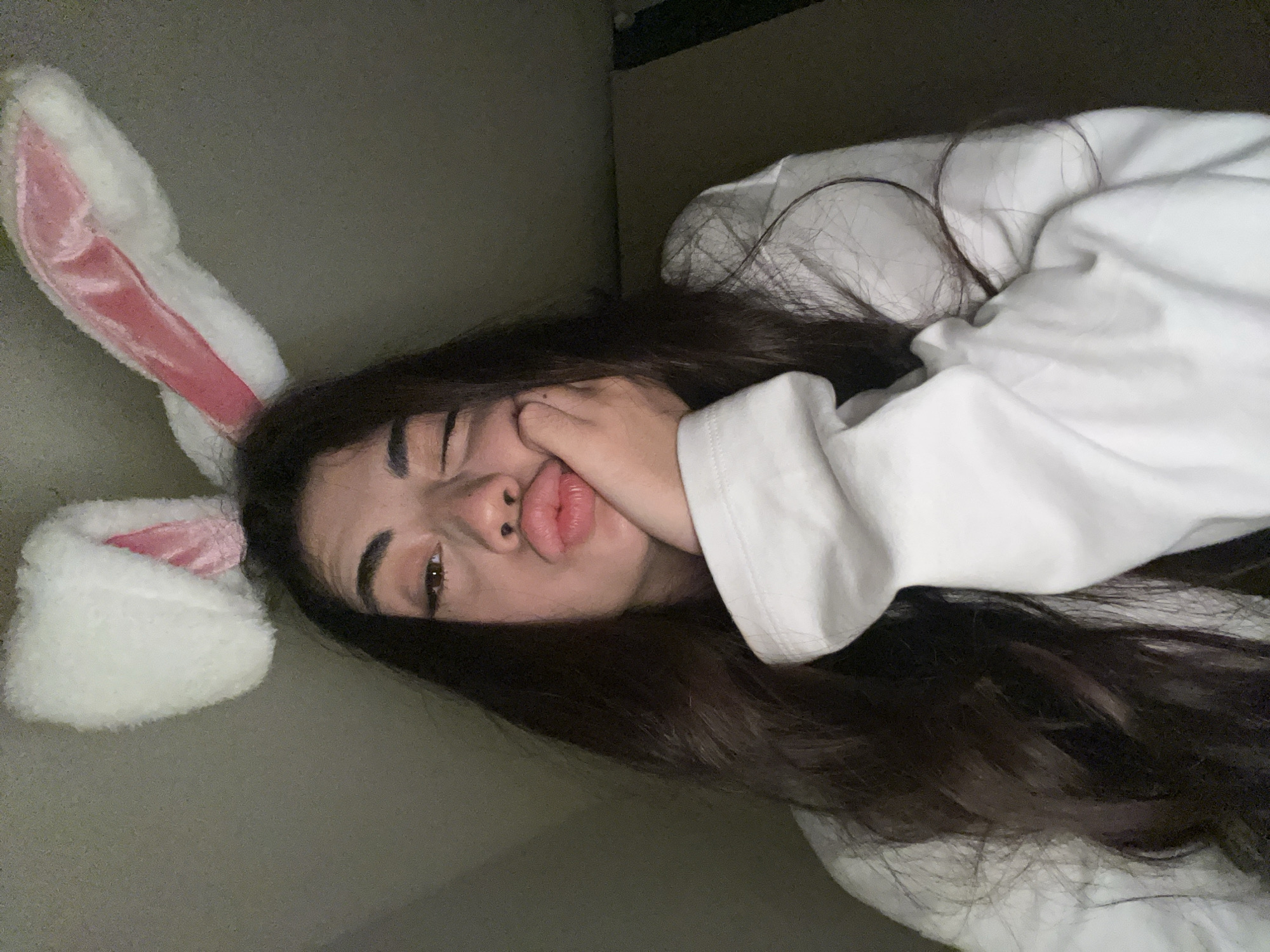 A young woman with long, dark hair and a white sweatshirt is looking down and to her left, winking with her right eye. She is wearing fuzzy white bunny ears with pink inner lining. Her left hand is supporting her chin, and her lips are pursed in a pout. The background appears to be an indoor space with textured walls and a ceiling that slopes downwards. The lighting is dim, casting shadows. There is no indication of the location being in Chotěšov, Czechia.