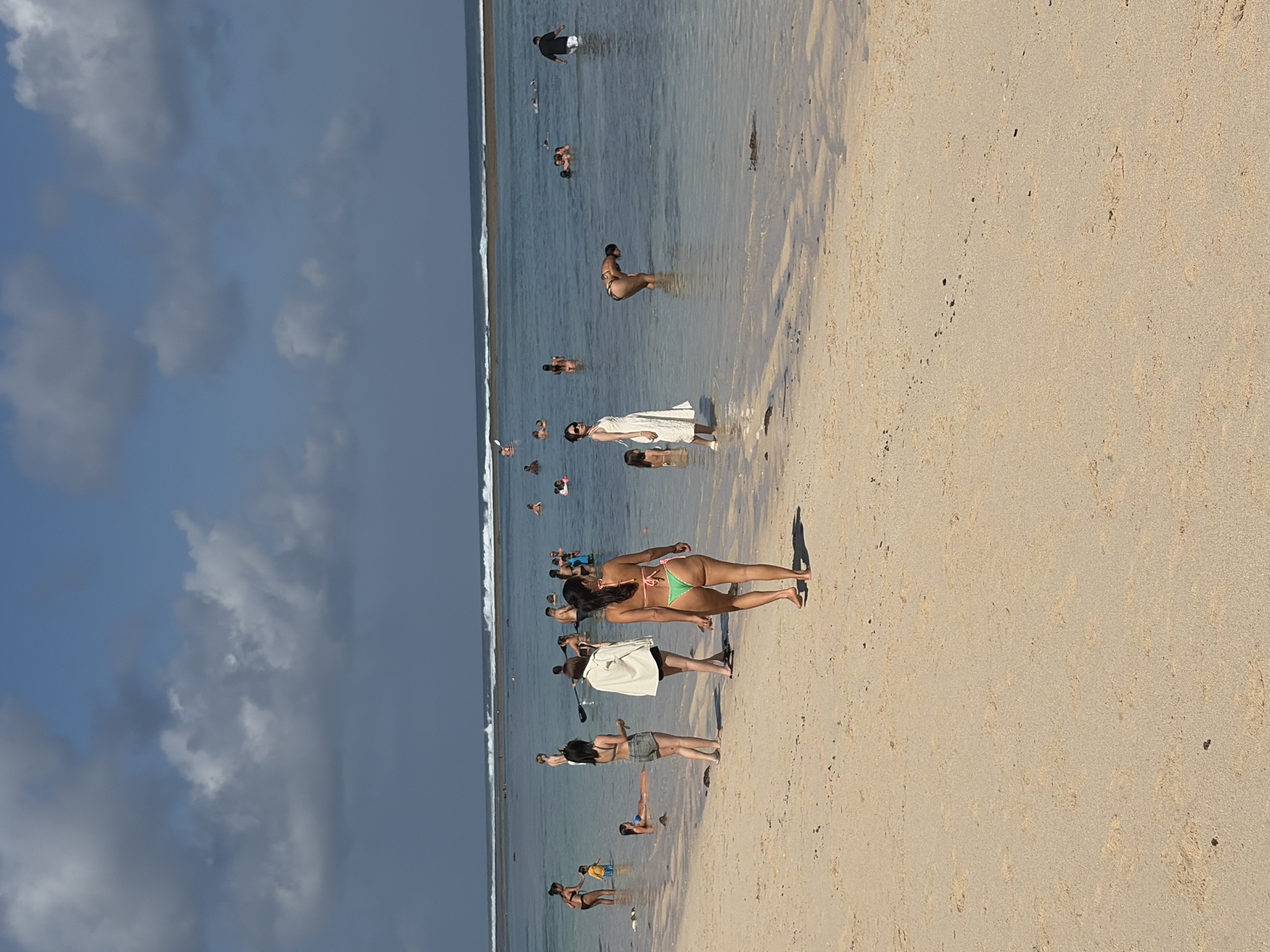 In Kuta Selatan, Indonesia, a sun-drenched beach scene unfolds with numerous people enjoying the shallow ocean waters and sandy shore under a partly cloudy sky. The water, a calm expanse of blue, laps gently onto the beach, creating a serene atmosphere. In the foreground, a woman in a green bikini walks away from the camera along the wet sand, her back to the viewer. Further into the water, a group of individuals are scattered, some wading, others swimming or standing. One woman in a white dress stands near the water's edge on the left side of the frame, looking towards the sea. Another woman in a bikini bends down in the water, seemingly observing something on the seabed. The beach itself is composed of light-colored sand, marked with subtle ripples from the receding tide. The distant horizon is marked by a line of breaking waves, suggesting a more active surf further out. The overall impression is one of a leisurely day at the beach, with various individuals engaged in typical seaside activities.