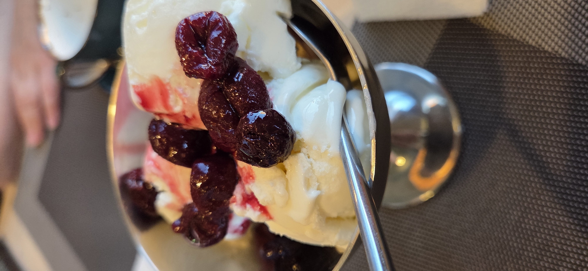 The scene captures a close-up, vertical perspective of a dessert served in a dining setting within Lipetsk, Russia. The primary subject is a metal, reflective silver-toned dessert bowl, elevated on a pedestal base, resting on a dark grey textured placemat or tablecloth. The bowl contains several scoops of white or off-white ice cream, extensively garnished with dark red to purple whole berries, likely cherries, and a visible reddish syrup partially mixed into the ice cream. A silver metallic spoon is partially submerged in the ice cream, extending towards the lower left of the frame. In the blurred background, a human hand is indistinctly visible, along with the partial view of a white ceramic cup featuring a golden rim.