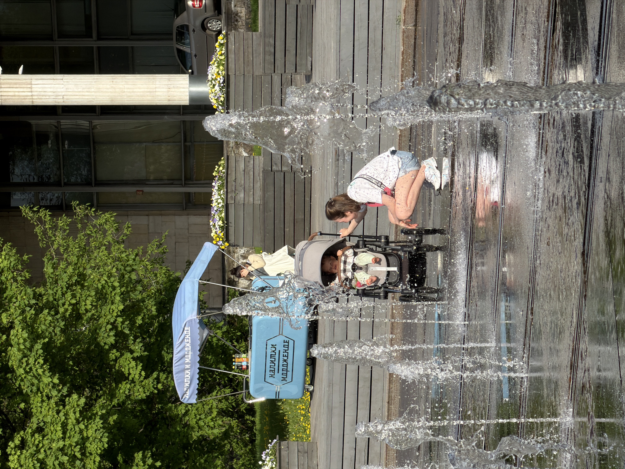 A woman crouches by a water fountain holding a stroller, near a blue beverage stand. The image shows a street scene in the city of Moscow, Russia. The fountain has multiple streams of water shooting upwards. The woman is wearing a white shirt and denim shorts. The stroller contains a baby. The blue beverage stand is advertising drinks and ice cream in Russian. There are green trees visible in the background, along with a building and a parked car. The ground is made of wooden planks. The photograph appears to be taken during daylight hours.