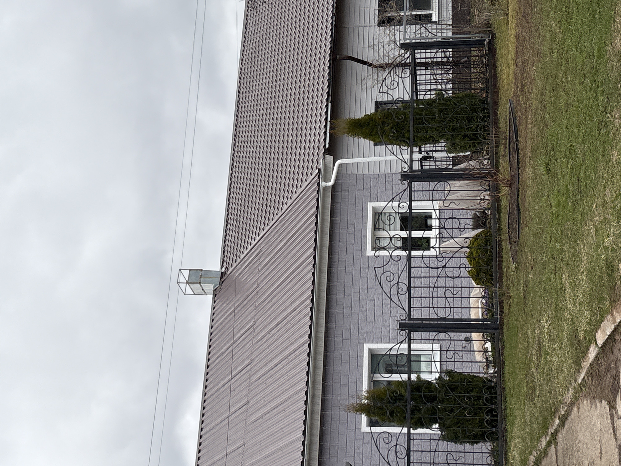 This image captures the exterior of a house in Прикамский, Russia, under an overcast sky. The house features a dark brown, corrugated metal tile roof with visible white gutters and a downspout. A metallic vent or chimney structure is positioned on the roof towards the left. The side of the house is clad in grey vertical siding, and two rectangular windows with white frames are visible.
In front of the house, a black wrought iron fence with intricate scrollwork and grid patterns provides a decorative boundary. Behind the fence, small, cone-shaped evergreen shrubs are planted, positioned in front of each window.
The ground consists of a lawn with green grass, interspersed with some patches of bare earth, particularly noticeable in the foreground. A section of a stone or concrete path is visible along the right edge of the frame, bordering the grass. Thin power lines are visible against the light grey, cloudy sky. No people or activities are present, and there is no visible text within the image. The scene suggests a quiet, possibly suburban or rural setting.
