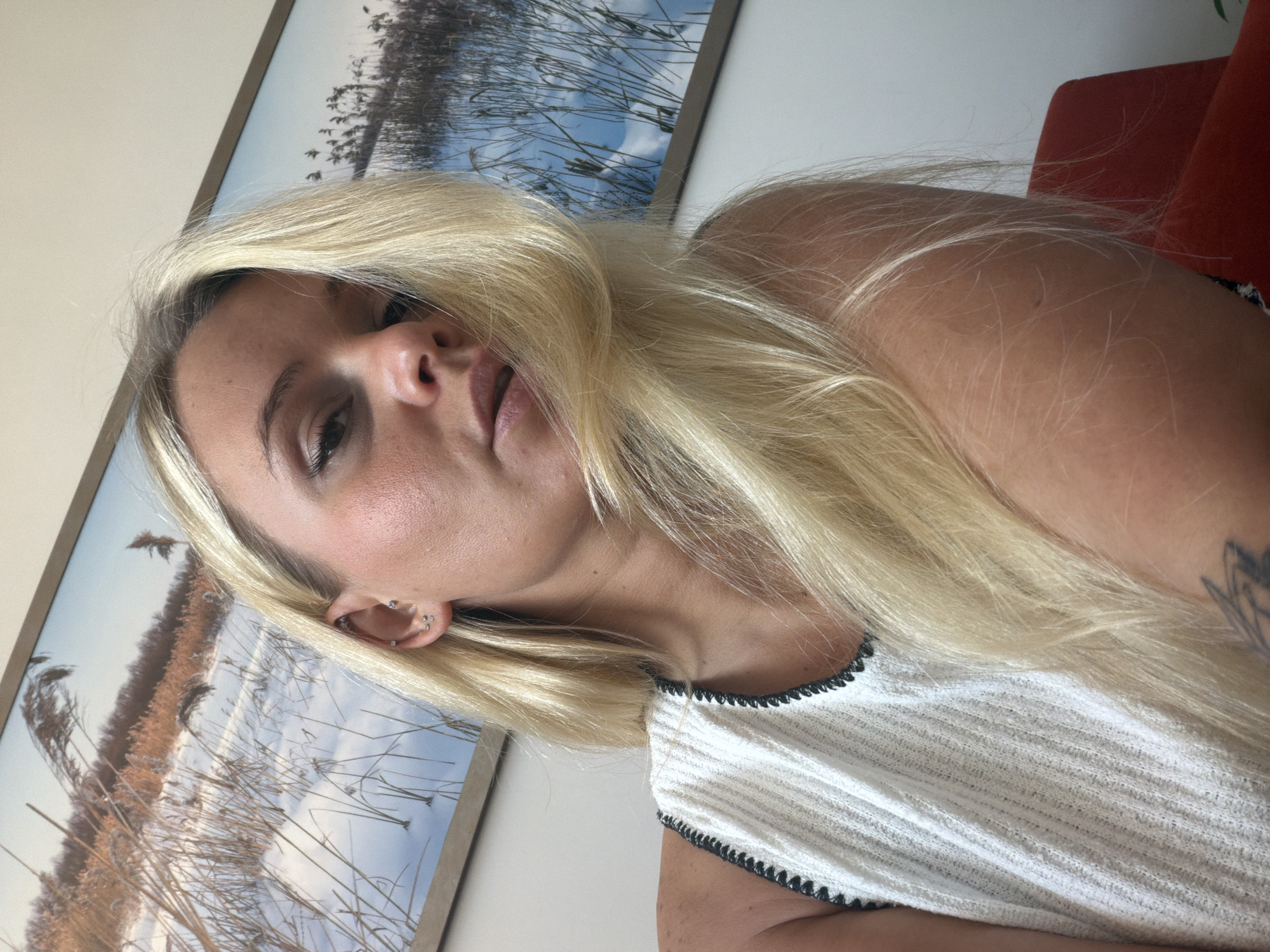 A blonde woman is captured in a close-up, selfie-style shot, her head tilted slightly towards the camera. She is wearing a white, sleeveless top with a black trim. Her blonde hair is predominantly in the foreground, obscuring part of her face and shoulder. In the background, a framed landscape artwork depicting a snowy scene with tall reeds is visible. The lighting suggests an indoor environment, possibly a home or office. The image does not contain any elements that would indicate its location in Sarapul, Russia.