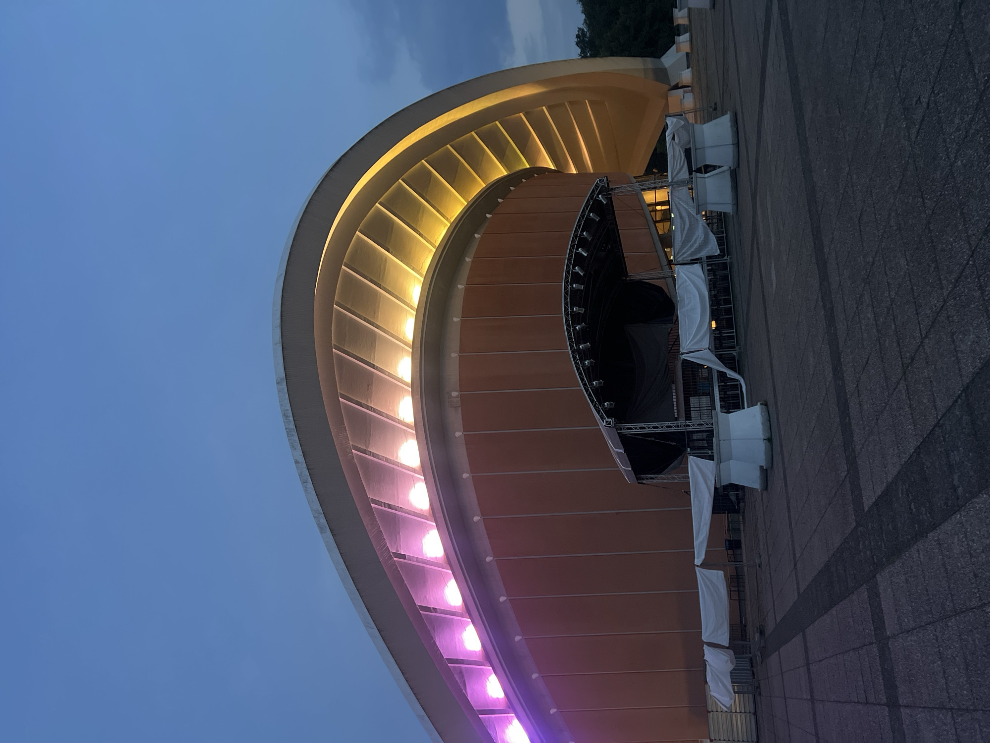 The image shows the exterior of the Philharmonie concert hall in Berlin, Germany, at dusk. The iconic curved roof of the building is illuminated with purple and yellow lights. In front of the building, a temporary stage setup with a black backdrop is visible, along with several white tent-like structures and white barriers. The ground is paved with large, dark grey tiles. The sky is a deep blue, indicative of the twilight hours. Trees are visible in the background to the right. There are no people in the frame.