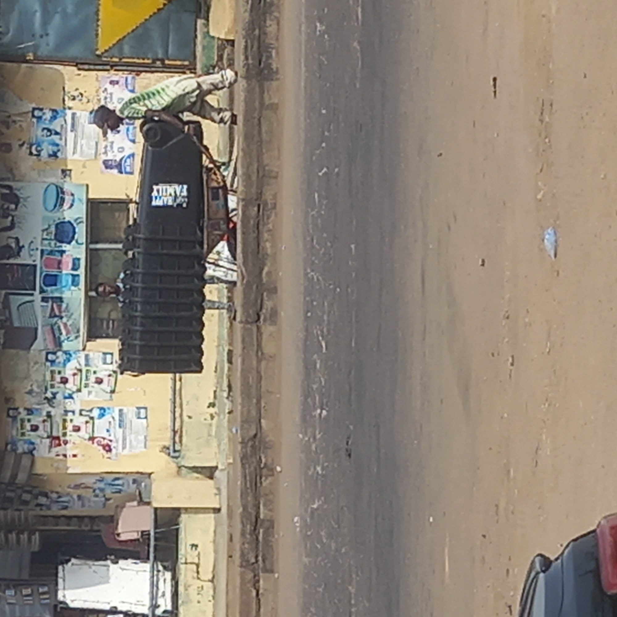 This image depicts a bustling street scene in what appears to be a market or commercial area.
In the foreground, on the left, is a building or shop front with a light-colored facade. Its walls are extensively covered with numerous posters and flyers, many displaying images of household items like buckets. A blue and yellow geometric pattern is visible on the upper part of the building. Through a window in the shop, the face of a person can be seen looking out. Various goods are displayed or stacked near the bottom of the building.
A man, wearing a light-colored striped shirt, trousers, and a cap, is positioned on the sidewalk or shoulder directly in front of the building. He is leaning over a large, black, ribbed container, possibly a water tank or storage bin. The container has white text printed vertically on its side, clearly reading "HAPPY FAMILY". The man appears to be interacting with or attending to the container.
To the right of the man and the building is a wide, unpaved or dirt road with a light brown, dusty surface. The front section of a dark-colored vehicle, featuring a red element that could be a taillight or reflector, is visible in the bottom right corner, suggesting it is parked or moving on the road.
The scene is brightly illuminated by natural light, indicating it is daytime. The overall impression is that of a lively street in a community setting, consistent with a location like Dadi Riba, Nigeria.
