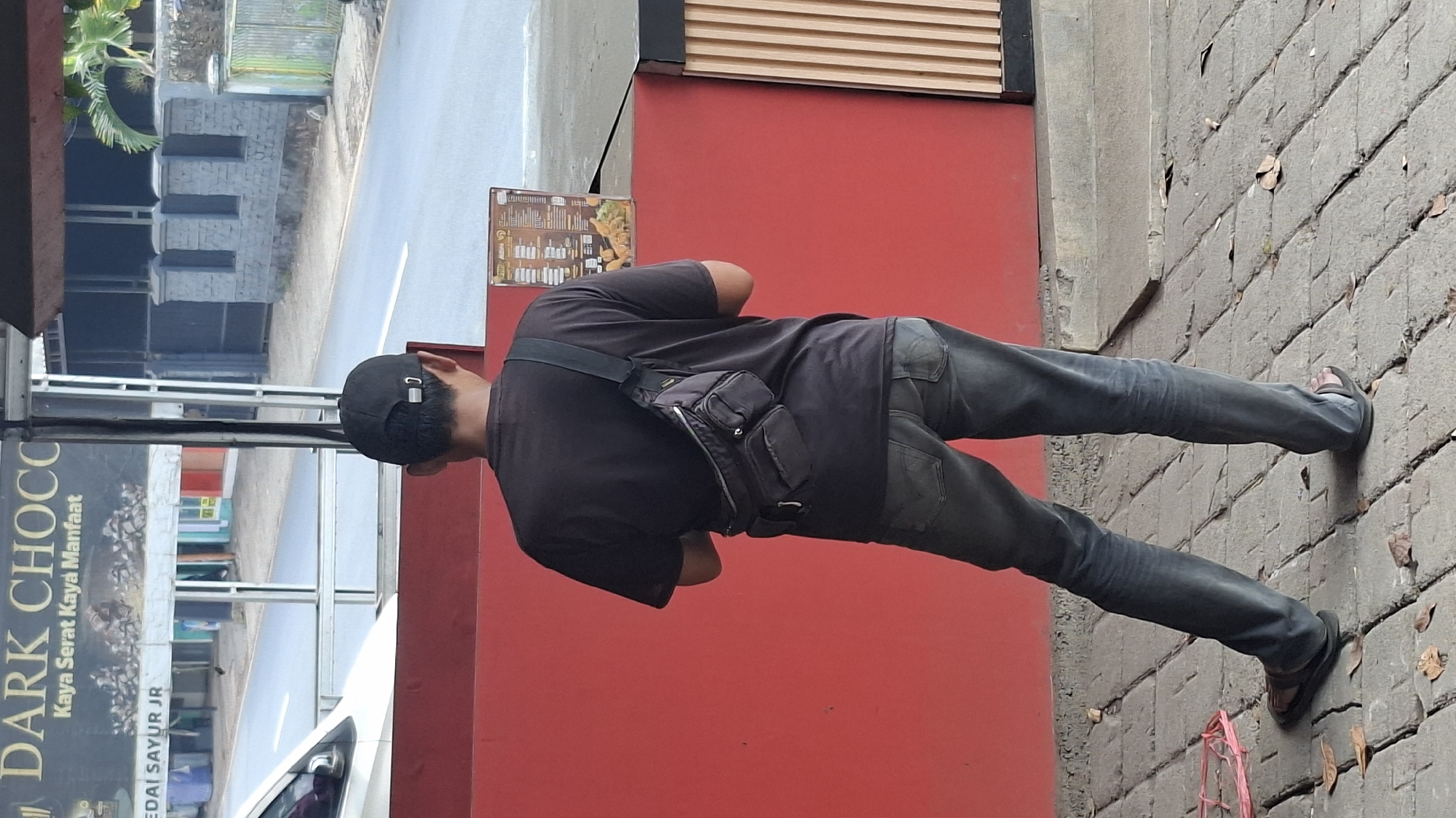 A person wearing a black t-shirt, dark denim jeans, and black flip-flops stands facing away from the camera. A black fanny pack is worn across their chest. The person is standing on a grey brick walkway in front of a bright red wall. Behind the person, a menu board is visible. The background includes a street with a white car, a dark building, and a stone fence. The scene appears to be located in Cilaku, Indonesia, as indicated by the signs with Indonesian text.