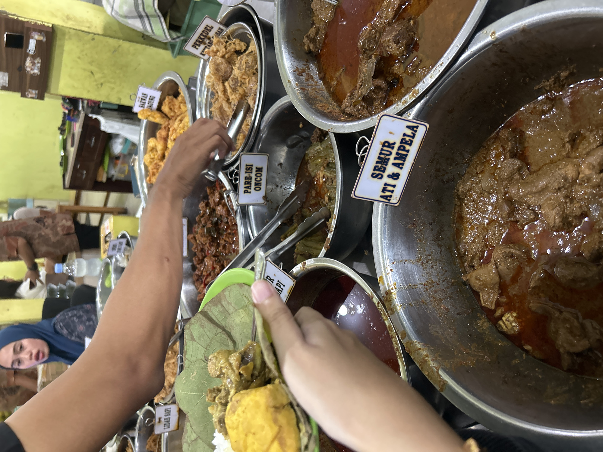 In a bustling Indonesian food stall in Cirebon, a person's arm is visible in the foreground, holding a plate of food. The plate contains rice, a yellow piece of food, and several pieces of meat with a sauce. The background shows a variety of food dishes in metal containers, including what appears to be beef rendang, semur hati & ampela (stewed liver and gizzard), pare-isi oncom (bitter melon with fermented soybean), bakwan jagung (corn fritters), and perkedel kentang (potato fritters). A woman wearing a blue hijab is visible in the middle ground, looking towards the food. The stall is brightly lit, with green walls and wooden furniture. The atmosphere is lively and suggestive of a popular eatery.