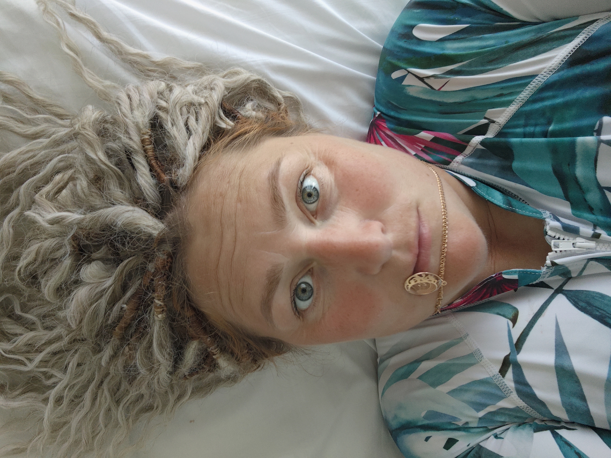 A close-up, overhead shot captures a woman lying on a white, textured surface, possibly a bed or couch. She has light-colored, wavy dreadlocks spread across the surface and around her head. Her face is turned towards the camera, with her left eye clearly visible and open, showing a blue iris. Her skin is fair with a rosy flush on her cheeks. She is wearing a zippered top with a tropical leaf print in shades of green, blue, and pink. A thin gold chain with a circular pendant rests on her collarbone. The image is devoid of any recognizable landmarks or context to indicate a specific location within the city of Sochi, Russia.