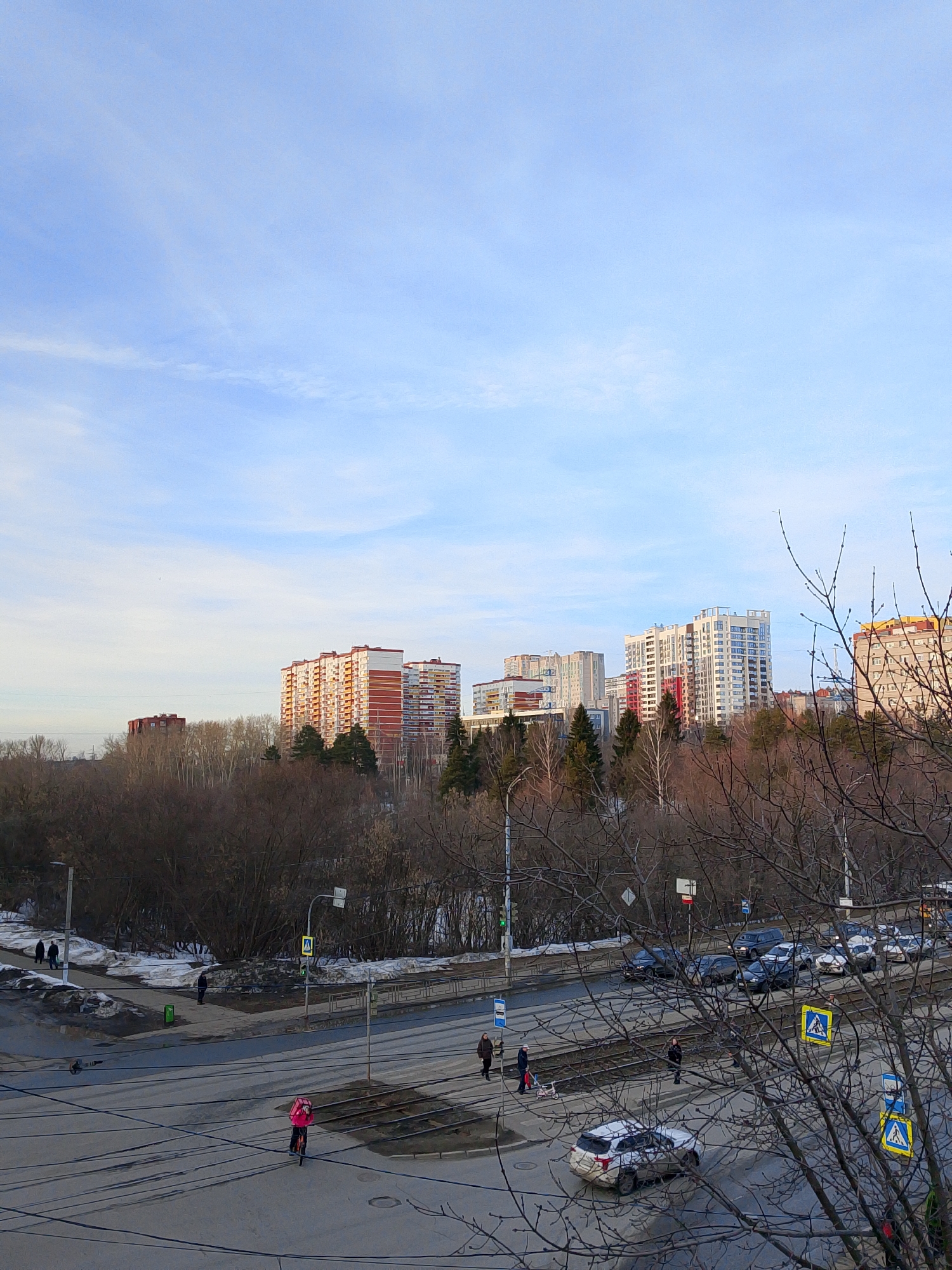 An elevated, wide-angle view depicts a multi-lane urban street scene in Izhevsk, Russia, under a clear light-blue sky with minimal cirrus clouds. The environment suggests late winter or early spring, characterized by bare deciduous trees and patches of melting, soiled snow and slush along pathways and unpaved areas.
In the mid-ground, a dense line of mostly bare trees and some scattered evergreens forms a natural barrier separating the street level from several distant multi-story residential buildings. These buildings vary in design, featuring facades with combinations of white, red-brown, yellow, and blue elements.
The foreground is dominated by a wide asphalt road, which appears wet or damp in sections. Running parallel and occasionally intersecting the road are twin tram tracks. Numerous utility poles with extensive overhead electrical wires crisscross the scene, particularly in the lower left quadrant.
Pedestrian crosswalks are marked by yellow signs featuring a walking figure. Multiple vehicles are present: several dark-colored cars are either parked or in motion along the street, and a light-colored SUV, visibly soiled with dirt or salt spray on its rear, is positioned near the tram tracks in the lower-center.
Several individuals are visible. On the road, near the tram tracks, a person wearing a bright pink jacket and a matching pink delivery backpack is riding a scooter. Other pedestrians are observed walking on sidewalks, with one individual pushing a stroller near a crosswalk on the right side of the frame. A green waste bin is visible on a sidewalk to the left. Bare tree branches from a large deciduous tree partially obscure the lower right portion of the scene.