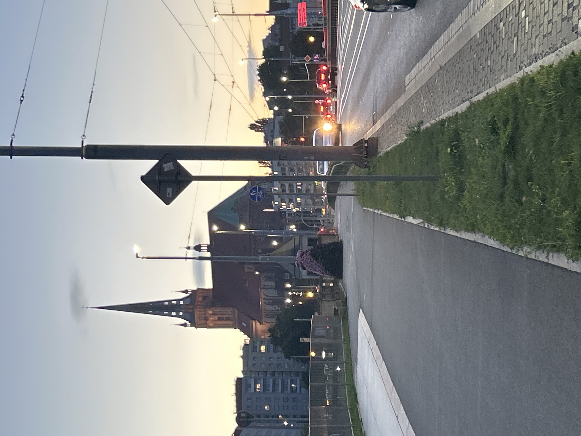 This image depicts a street scene at dusk in the city of Szczecin, Poland, specifically near the St. James Cathedral. In the background, the distinct spired architecture of the St. James Cathedral is visible, illuminated by the fading daylight. Adjacent to the cathedral, a modern building with multiple floors and lit windows is present, suggesting a mixed urban environment.
The foreground features a paved pedestrian walkway and a road with several vehicles, including cars and possibly a tram, indicated by overhead electrical wires and traffic signals. The road surface is asphalt, with cobblestone and concrete bordering the pedestrian areas. Streetlights are on, casting a warm glow on the surroundings. A prominent lamppost with signage is positioned near the center of the frame.
A section of a metal fence runs along the left side of the walkway, separating it from a lower level, possibly a park or a riverbank, where a dense, purplish bush or flower arrangement is situated. The sky above is a pale gradient of blue and orange, typical of sunset. The overall impression is of a calm, urban evening.