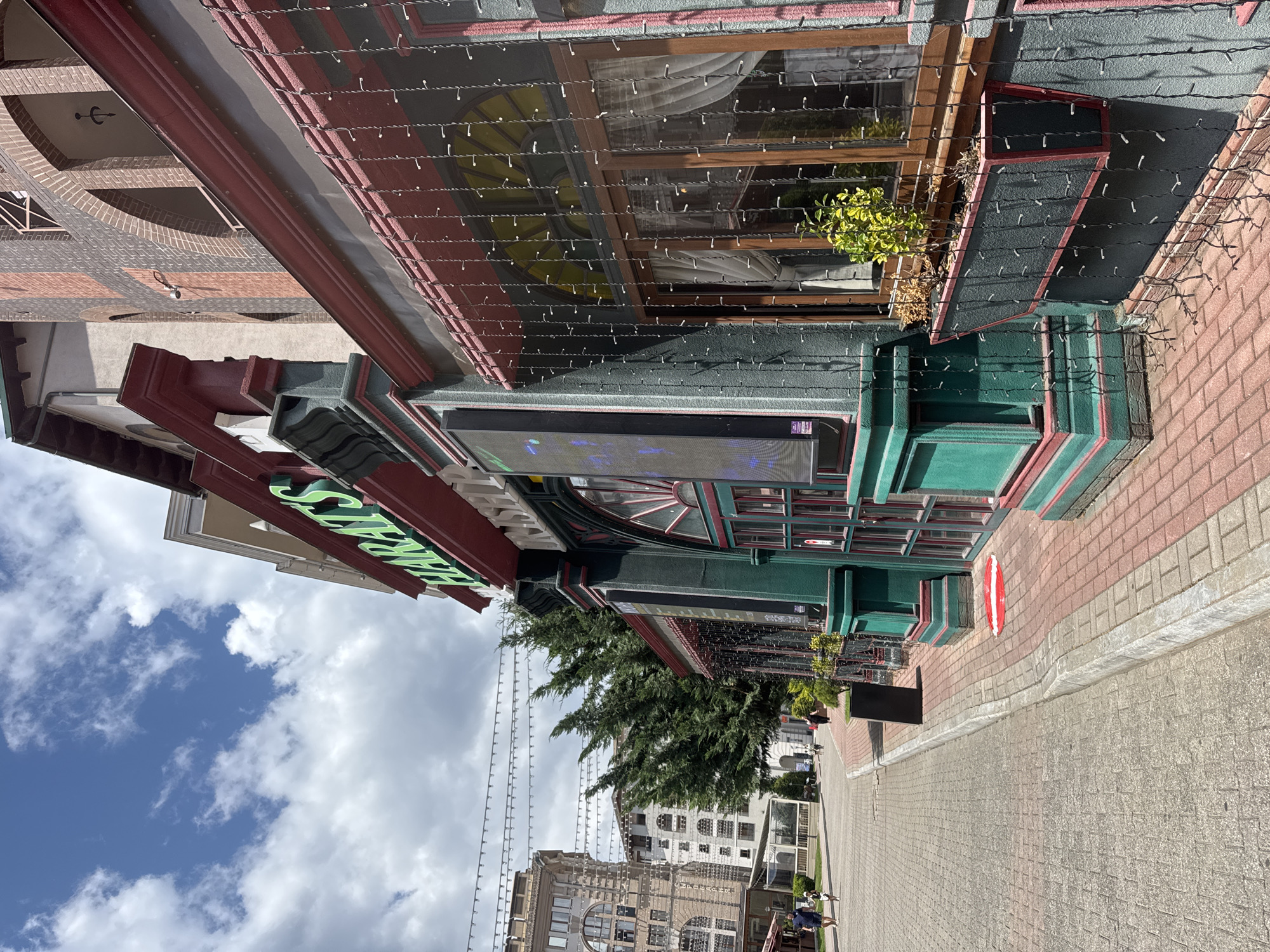 This is a ground-level, eye-level, slightly angled exterior shot of the HARATS Irish Pub in Estosadok, Russia, on a bright, cloudy day. The pub has a distinctive green and red facade, with a sign featuring green lettering that reads "HARATS". String lights, possibly for decoration, hang down from an awning above a window. A planter box with a small green plant is mounted on the wall next to the window. The pavement in front of the pub is made of red and grey bricks. In the background, several large buildings, likely hotels or apartments, can be seen, along with people walking on the sidewalk and trees lining the street. The sky is blue with scattered white clouds.