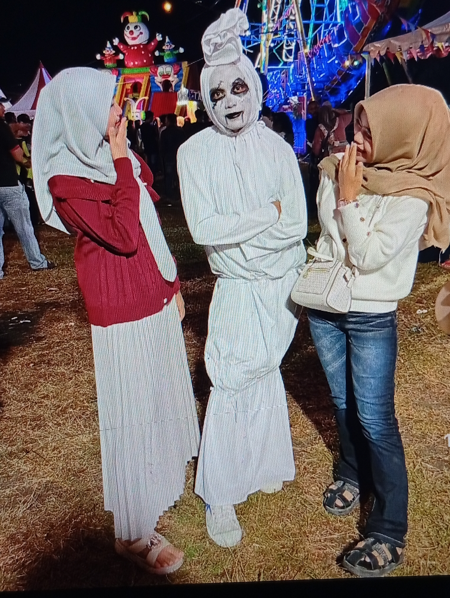 This image captures a nighttime scene at an outdoor carnival or fair in Lhokseumawe, Indonesia. The ground beneath the subjects is covered in dry, yellowish-brown grass.
In the foreground, three individuals are positioned facing the camera.
1. **Left Individual:** A person, likely female, wears a light grey hijab, a long-sleeved red jacket or top over a white garment, and a long white skirt. Her right hand is raised to her mouth, suggesting amusement or surprise. She wears light-colored flat sandals.
2. **Center Individual:** A person is dressed in an all-white, loose-fitting costume that covers the entire body and head. The face is painted white with prominent dark rings around the eyes, dark vertical lines extending below the eyes, and a dark, wide mouth line. The head covering is gathered at the top, resembling a traditional Indonesian pocong costume. This individual's arms are crossed at the chest, and they wear white, closed-toe footwear.
3. **Right Individual:** A person, likely female, wears a light tan hijab, a white long-sleeved sweater, and blue denim jeans. Her hands are clasped together in front of her chest, also suggesting amusement or shyness. She carries a small, light-colored structured handbag with a textured surface, possibly woven, slung over her left shoulder. She wears dark multi-strap sandals.
The background features illuminated carnival attractions. To the left of the center, a large inflatable structure depicting a red clown with smaller clowns is visible atop a colorful base. To the right, a tall structure adorned with blue and multicolored lights, consistent with a Ferris wheel or similar ride, dominates the skyline. A white tent with decorative colorful pennants is also partially visible behind the right individual. Artificial lighting from the attractions illuminates the scene against the dark night sky. The overall atmosphere appears festive and lighthearted, with the individuals seemingly posing for a photograph.