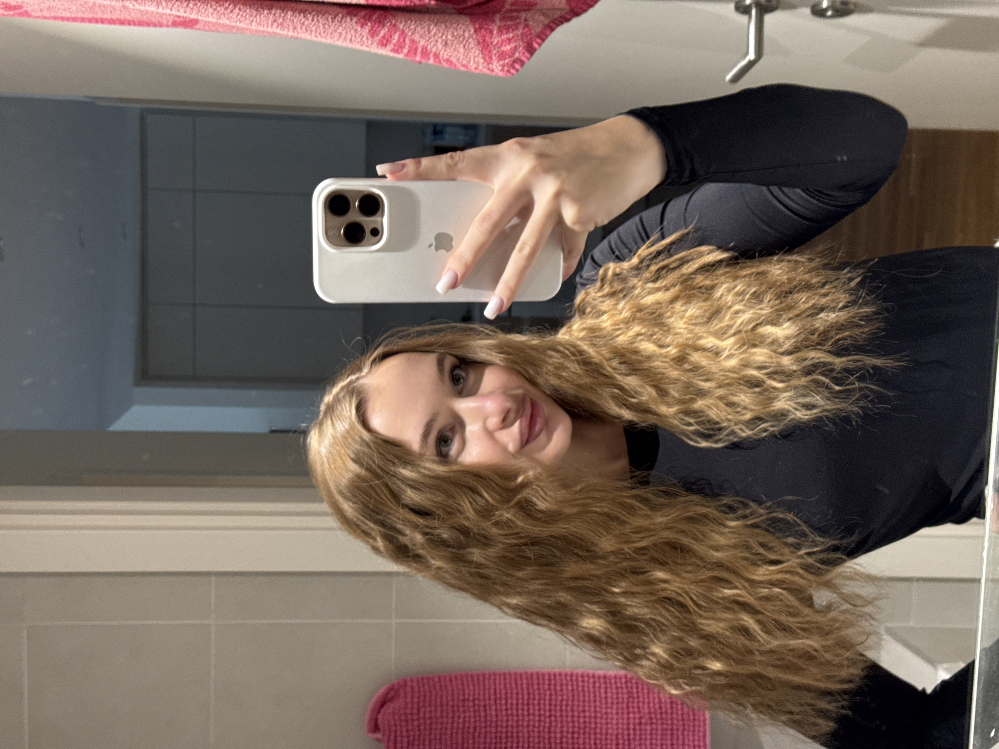 A young woman is taking a selfie in a bathroom using a white iPhone. She is wearing a long-sleeved black top. She has long, wavy blonde hair. The bathroom has a light-colored tile wall and a pink towel is visible. There is a pink bath mat on the floor. The image does not contain information to determine the location in Belgrade, Serbia.