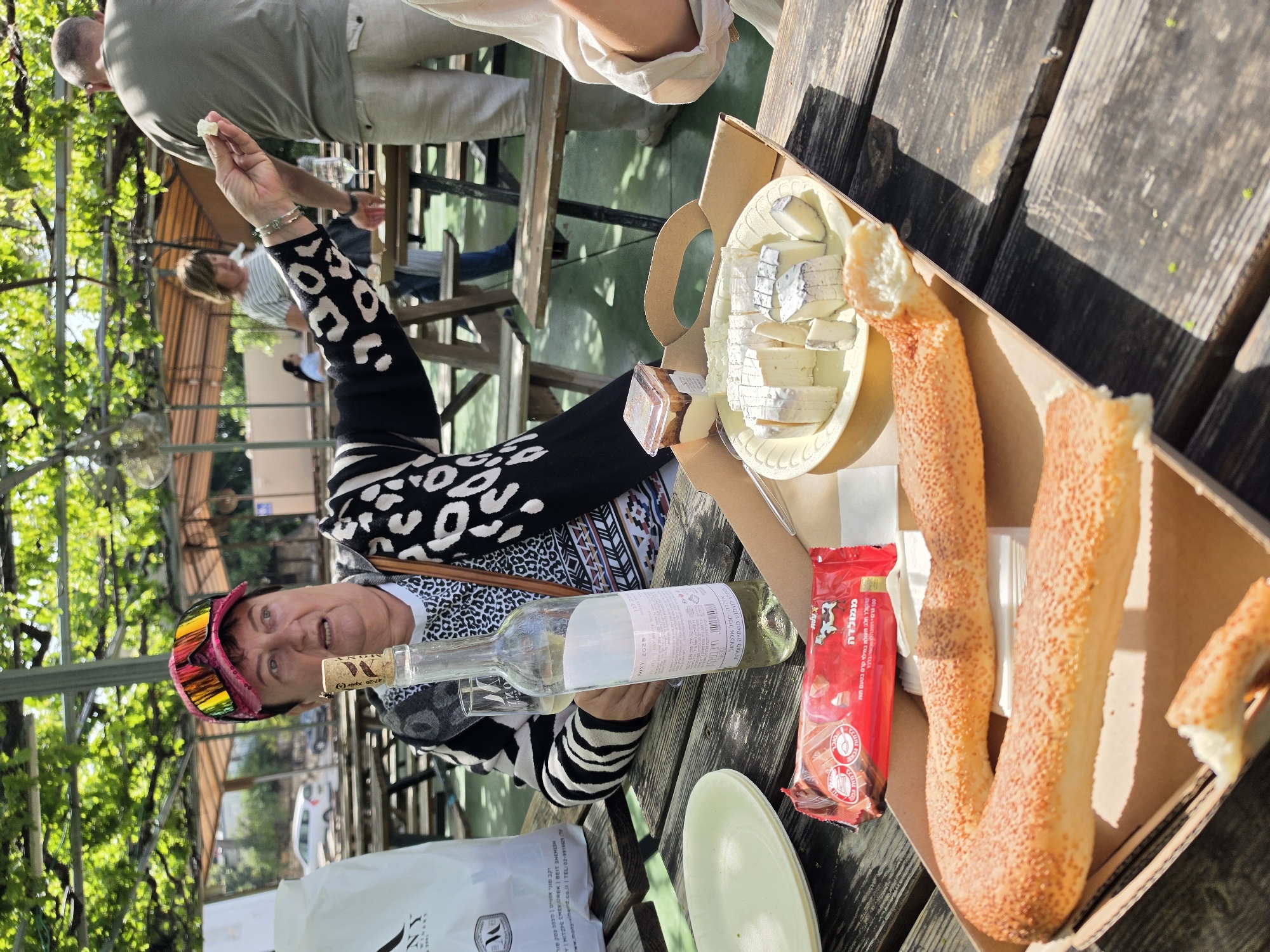 Here is a description of the image:
A woman sits at a wooden picnic table with food and drinks. She is wearing a black and white patterned sweater and a pair of brightly colored sunglasses perched on her forehead. She holds a small piece of food in her raised hand.
On the table are a bottle of wine, a platter of cheese, a package of chocolate, and twisted sesame seed bread in a cardboard container. There is a paper bag and a plate on the table, and an open bottle of wine is also visible.
The picnic table is located outdoors, under a canopy of greenery. The scene is in Mate Yehuda Regional Council, Israel, as inferred from the text on the bottle label. Other people are visible in the background, seated at similar tables.