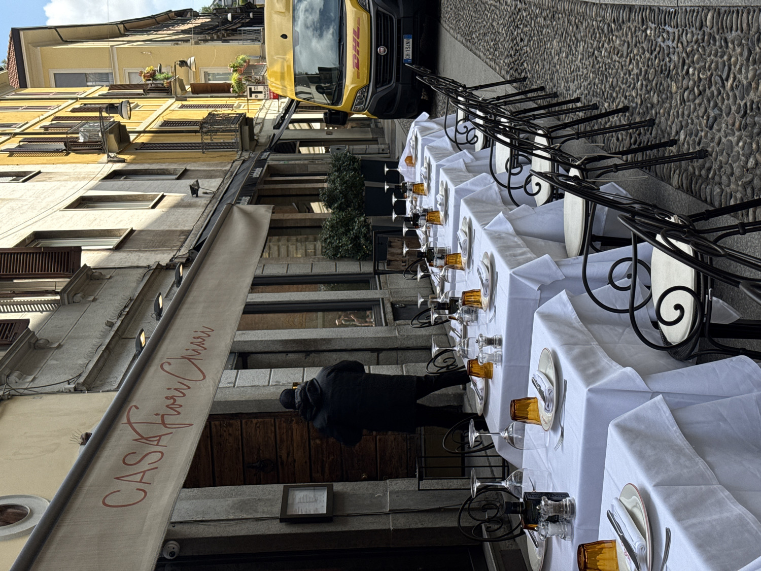 This image captures an outdoor dining setup along a cobblestone street in Milan, Italy, oriented vertically with buildings on the left and the street extending from foreground to background.
The scene is dominated by a long row of rectangular tables, meticulously covered with white tablecloths and set for dining. Each place setting includes a white plate, cutlery, a clear wine glass, and a distinctive amber-colored tumbler. Black metal chairs, featuring decorative scrollwork on their backs, are neatly arranged at the tables.
On the left side, a multi-story building with a light grey facade lines the street, featuring windows, some with closed wooden shutters. A large, beige awning extends from this building over the tables, displaying the red script text "CASA Fiori Chiari". Further down the street, another building with a bright yellow facade is visible, adorned with balconies and potted plants.
A single person, dressed in dark attire, stands near the entrance of the restaurant, their back to the camera, seemingly involved in preparations. In the distance, at the end of the cobblestone street, a yellow DHL delivery van is parked, its "DHL" logo clearly visible. The license plate "CH 12NH" is partially visible on the van.
The photograph depicts a daytime scene, likely under clear skies, given the bright and relatively even lighting, suggesting pleasant weather for outdoor dining. The atmosphere is quiet and anticipatory, with the restaurant prepared for service but currently devoid of customers. The cobblestone street and traditional architecture reinforce the Italian urban setting.