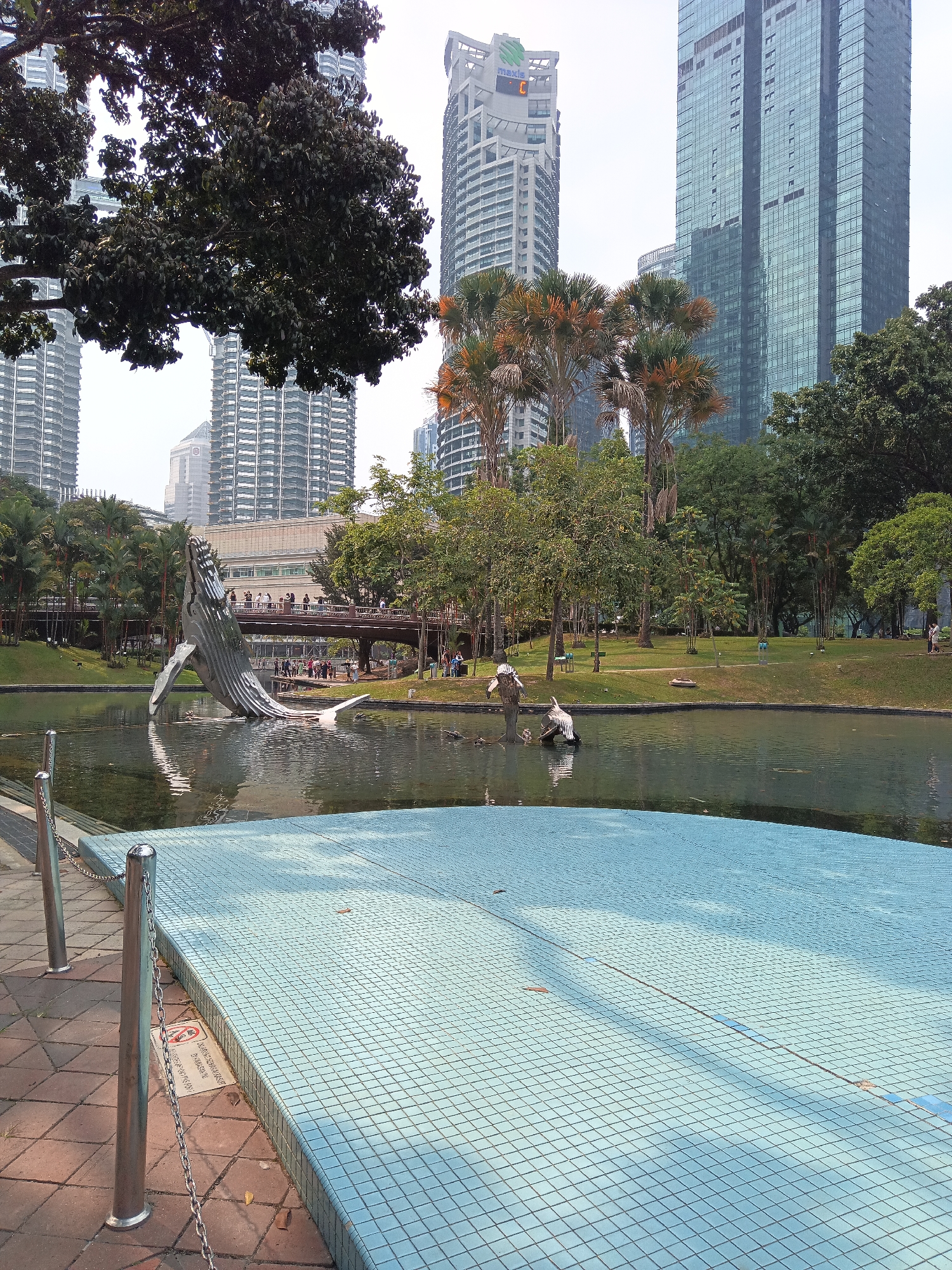 The image depicts a scenic view of a park in Kuala Lumpur, Malaysia, featuring a large whale sculpture partially submerged in a pond. In the background, modern skyscrapers, including the Maxis Tower with its distinctive green logo, dominate the skyline. Palm trees and lush greenery surround the pond, creating a natural oasis within the urban environment. Several people are visible walking along a bridge and in the park, enjoying the public space. The foreground shows a tiled area and a chain-link fence, suggesting this is a designed recreational area. The overall impression is one of a well-maintained park integrated into the bustling city.