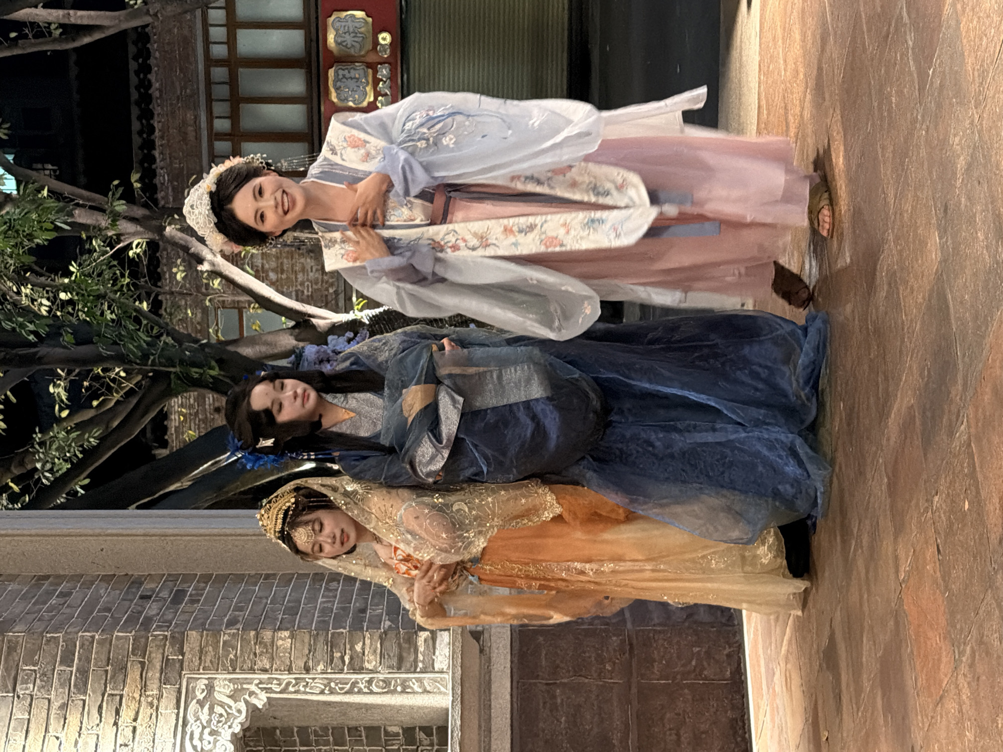 Three women in traditional Chinese attire are standing in what appears to be an outdoor public space at night. The woman on the left is wearing an orange and gold dress with a sheer, embroidered veil. The woman in the middle is in a deep blue garment, and the woman on the right is in a pink and light blue ensemble with intricate floral embroidery and a decorative headdress. They are standing on a paved surface in front of a brick building with ornate architectural details. Trees with visible foliage are behind them, suggesting an urban environment. The lighting indicates it is evening or nighttime. The woman on the right is smiling and has her hands on her chest, while the other two women have more neutral expressions. The scene is likely taking place in Chancheng District, China, given the context of traditional Chinese clothing and architecture.