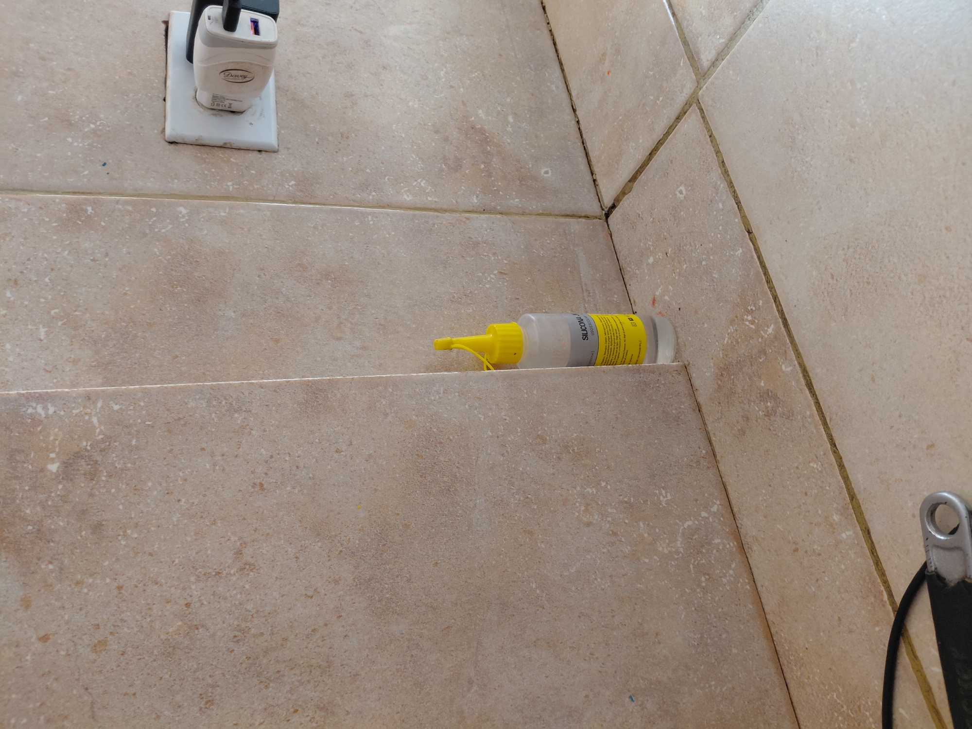 The image captures a close-up view of a tiled indoor surface, likely a floor or wall, in what appears to be a functional or utility area. The tiles are a light beige or sandy brown color with a mottled, speckled pattern, and dark grout lines separate them.
In the upper left quadrant, a white electrical outlet is visible, into which a white power adapter/charger is plugged. The adapter has "Devary" inscribed on its side, along with a "CE" mark and other smaller, unreadable text.
Towards the center-right of the image, lying horizontally on one of the tiled surfaces, is a translucent bottle with a bright yellow cap and nozzle. The bottle contains a clear or white substance. Visible text on the bottle's label includes "SILICONA" (Spanish for Silicone) and "PROFESSIONAL," with smaller, unreadable text below.
The perspective suggests the photo was taken from slightly above, looking down into a corner or step, as grout lines indicate distinct planes of tile converging. In the bottom right corner, a small portion of a metallic object with a black strap attached is partially visible.
No people are present, and no explicit activity is taking place; the scene is static. The items suggest a domestic or maintenance setting. The lighting is even, suggesting an indoor environment with either natural or artificial light. The overall impression is practical and functional. This scene is situated in Santa Ana, Venezuela.