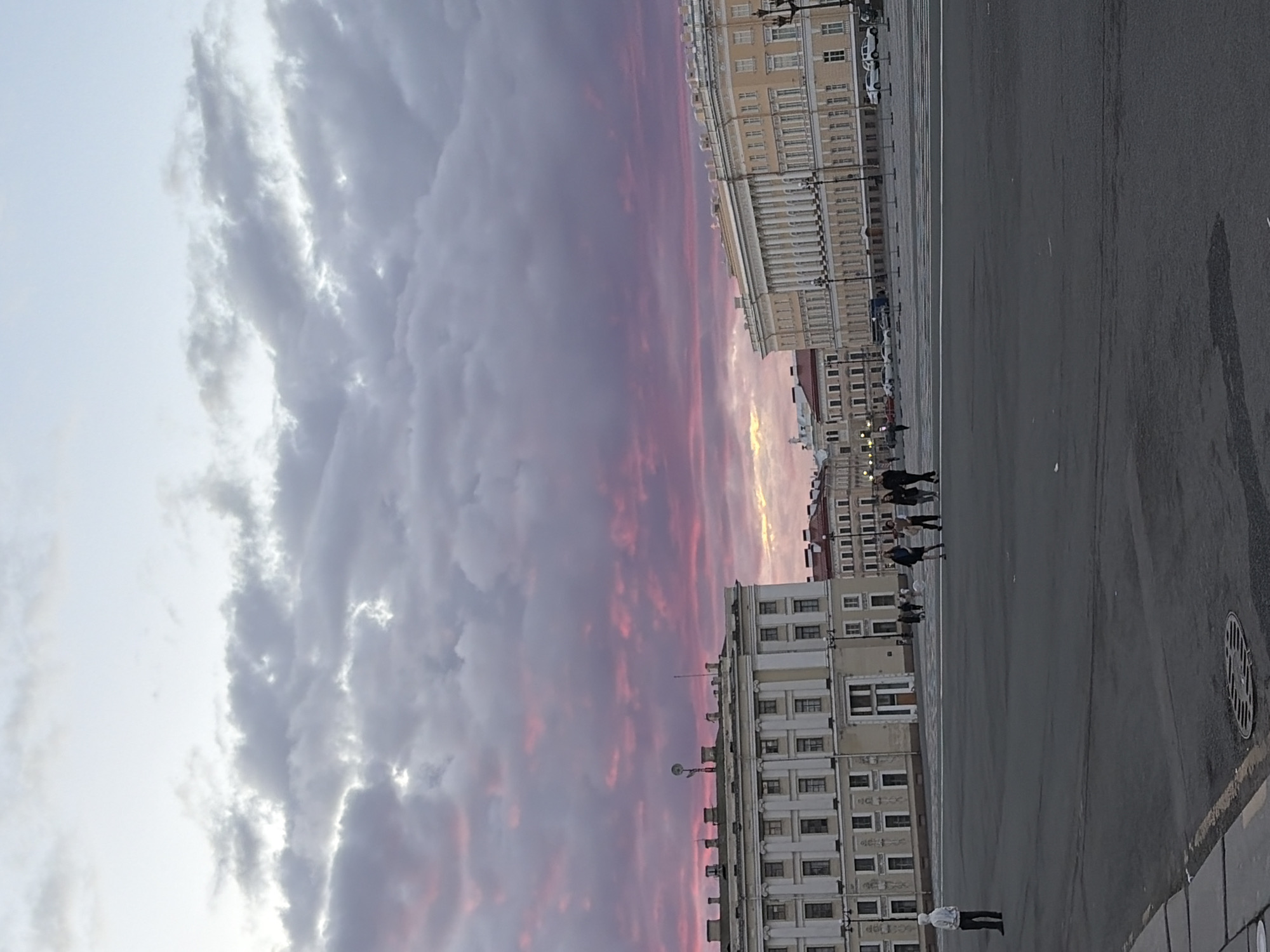 The image depicts Palace Square in Saint Petersburg, Russia, at sunset. The sky is filled with dramatic clouds tinged with pink and purple hues. Two grand, neoclassical buildings flank the expansive, paved square, with columns and numerous windows characteristic of the architecture. Several people are visible in the distance on the square, appearing to walk. One individual stands in the foreground on the left, facing away from the camera. The paved ground in the foreground shows some wet patches and a circular manhole cover. The overall scene suggests a calm, late evening in a historic urban setting.