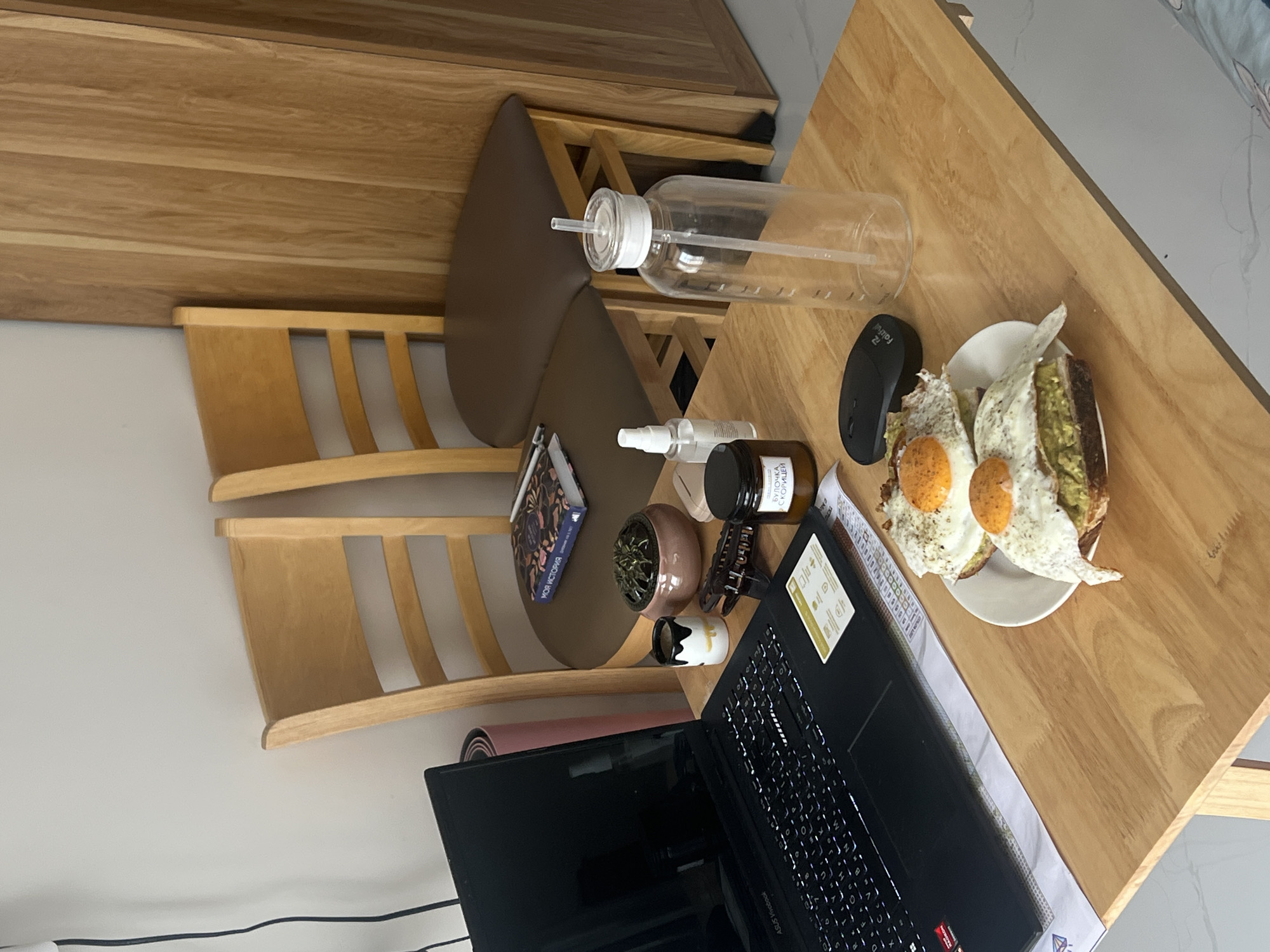 A slightly elevated, angled shot focuses on a wooden desk with breakfast, work, and personal items, likely indoors. On the desk is an open laptop with a black screen and visible keyboard, alongside a plate of food featuring fried eggs on toast. A black computer mouse rests near the plate. Other objects present on the desk include a clear water bottle, a small potted plant, a hair clip, a small decorative figure, and a cylindrical jar with text in a Cyrillic script. There is also a book placed between the desk and a wooden chair. Behind the chair, a pink yoga mat is partially visible. The image indicates a personal or work space. The location in the city of Nam Natural Heritage Area Trang, Vietnam, is not evident from the image, as there are no recognizable external environmental cues.