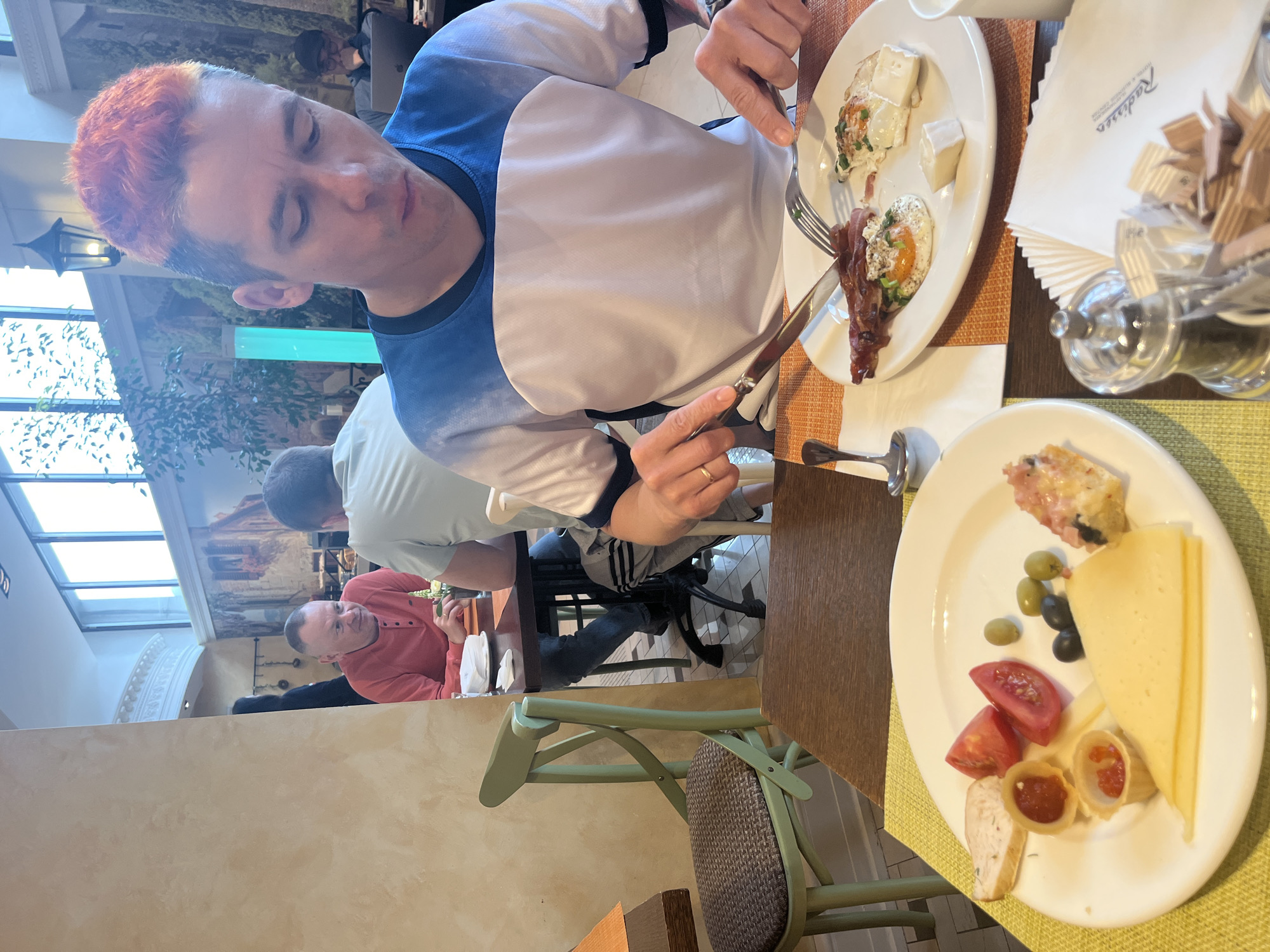 A restaurant scene depicts a man with reddish-orange hair at a table, eating from a white plate with bacon, eggs, and cheese, using a knife and fork. Another white plate contains sliced cheese, tomatoes, olives, and stuffed pasta shapes. The table is also set with a small container of condiments, a stack of napkins with "Radisson" printed on them, and a cup. Two other men are seated at a table in the background. The restaurant setting includes a high ceiling with windows and a decorative mural along the wall. The scene appears to be in the city of Moscow, Russia, given the Radisson napkins, suggesting the hotel chain's presence.
